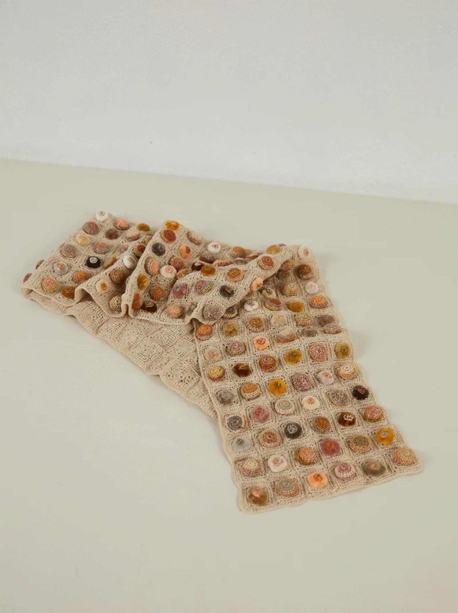 Sophie Digard knitted scarf on white surface. Grid pattern of multicoloured circles in earthy tones. Light-coloured background complements minimalist aesthetic.