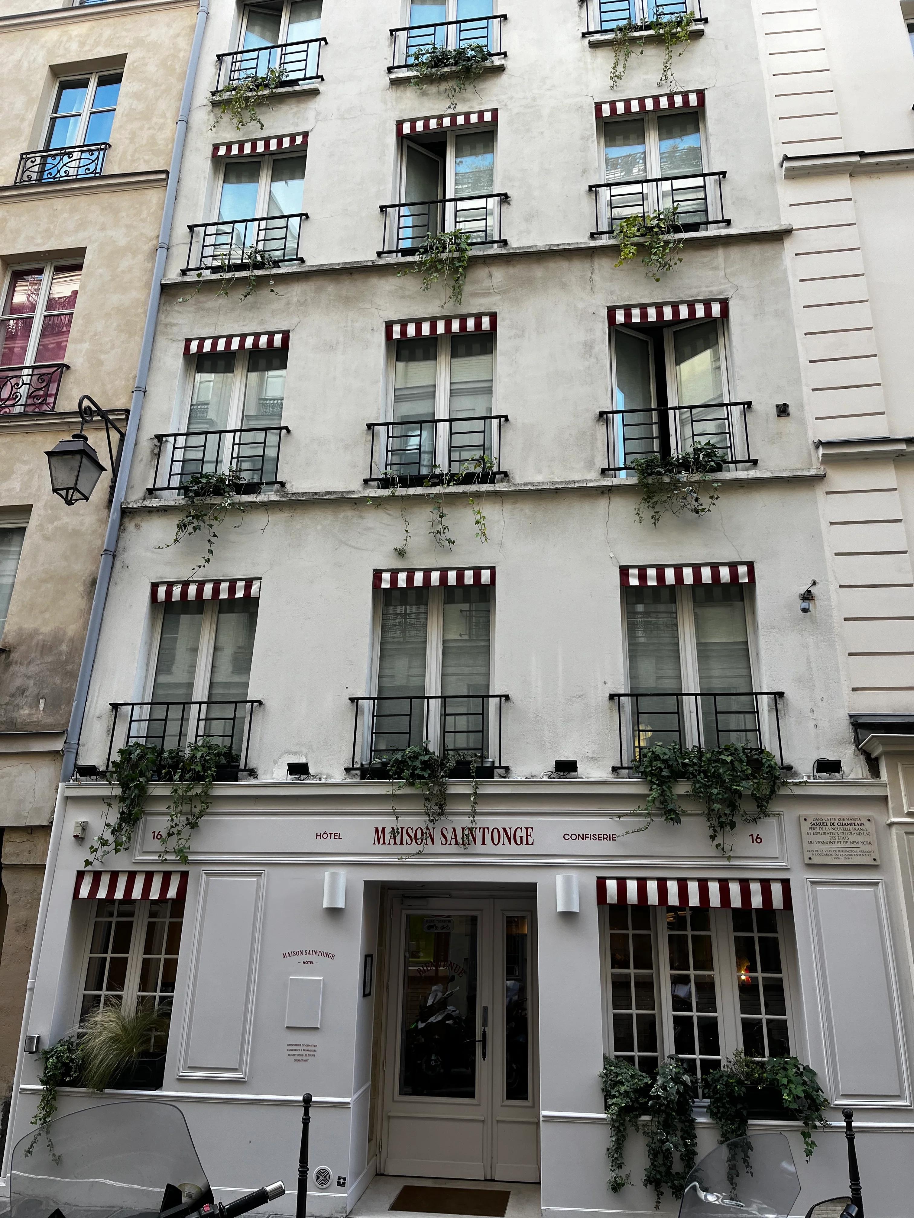 Five-story building with white and beige exterior. Windows with plants and striped awnings. "Maison Saintonge" sign above doorway.