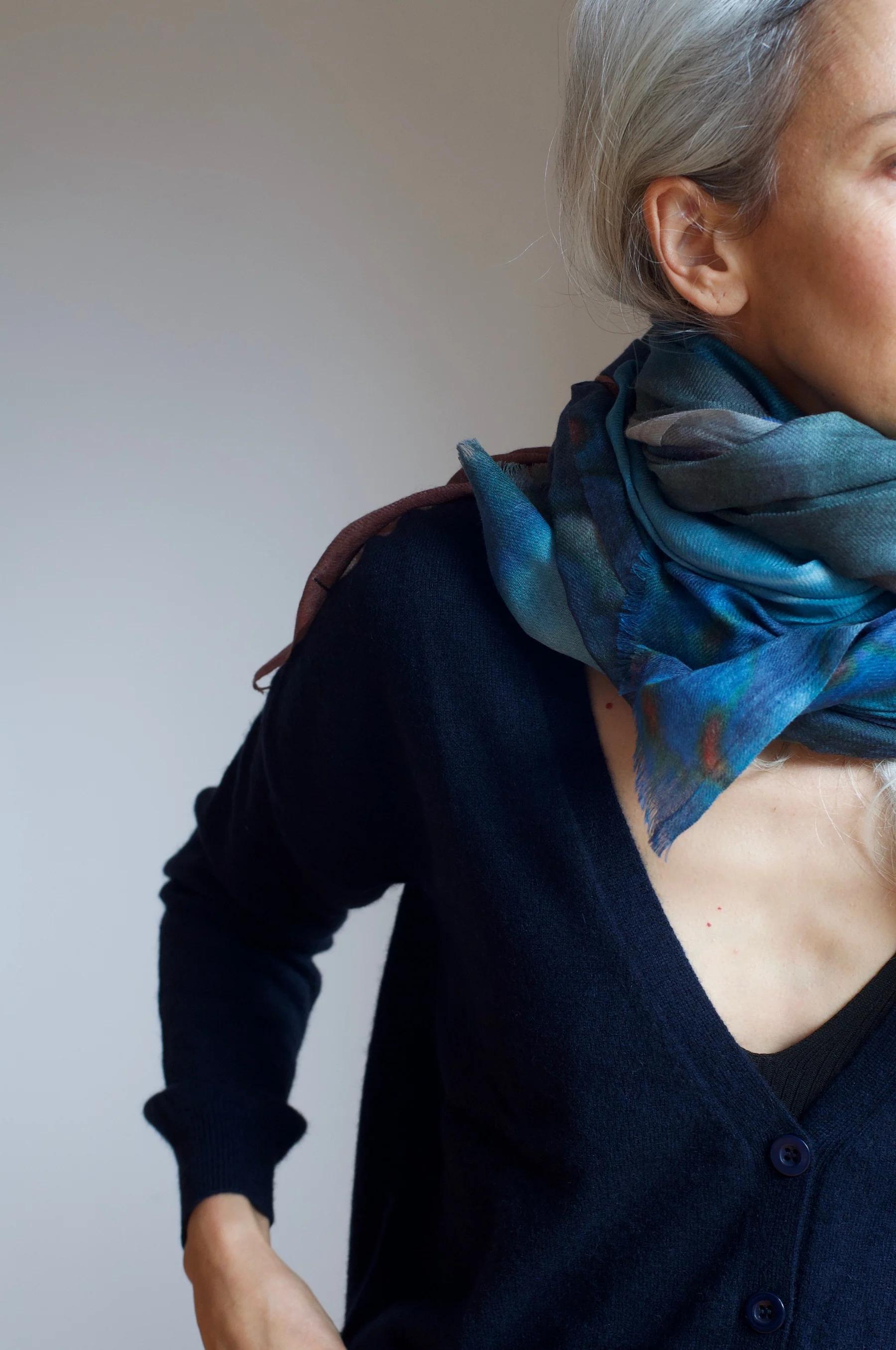 Side view of light-haired person in dark cardigan and blue Good & Co scarf. Plain grey background showcases autumnal loungewear.