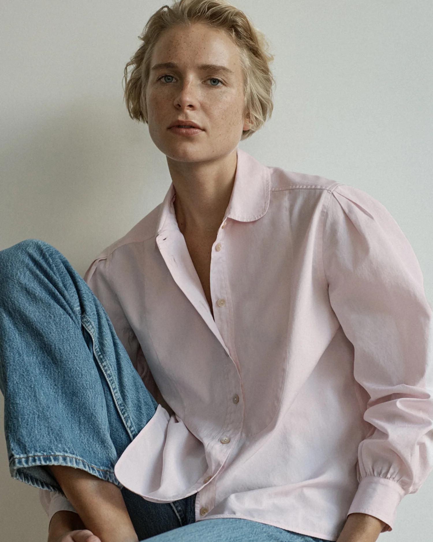 Person with short blonde hair in light pink button-up and jeans. Sitting with one knee bent. Plain backdrop.