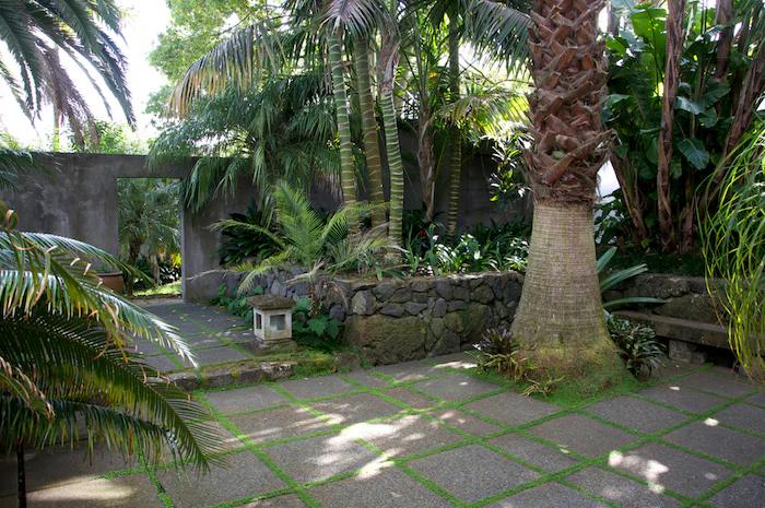 Tropical garden path with moss-lined stones. Stone lantern by Rachel Carley centres the scene. Doorway filters light, creating secluded ambiance.