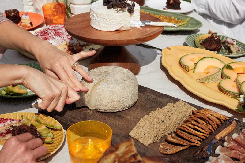 A person slices cheese on a wooden board at a garden table filled with crackers, fruit, and snacks. Guests reach for food during a summer lunch.