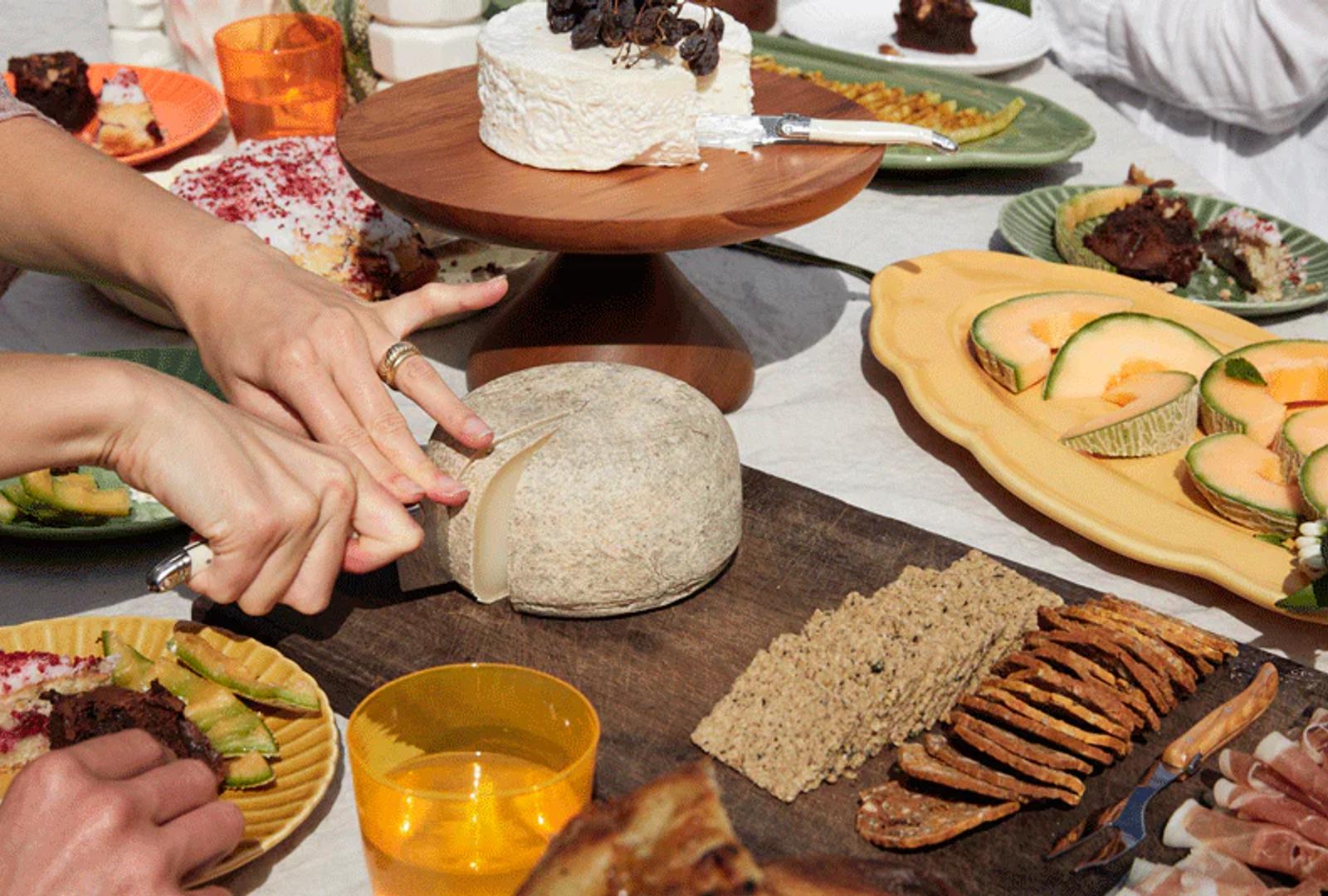 A person slices cheese on a wooden board at a garden table filled with crackers, fruit, and snacks. Guests reach for food during a summer lunch.