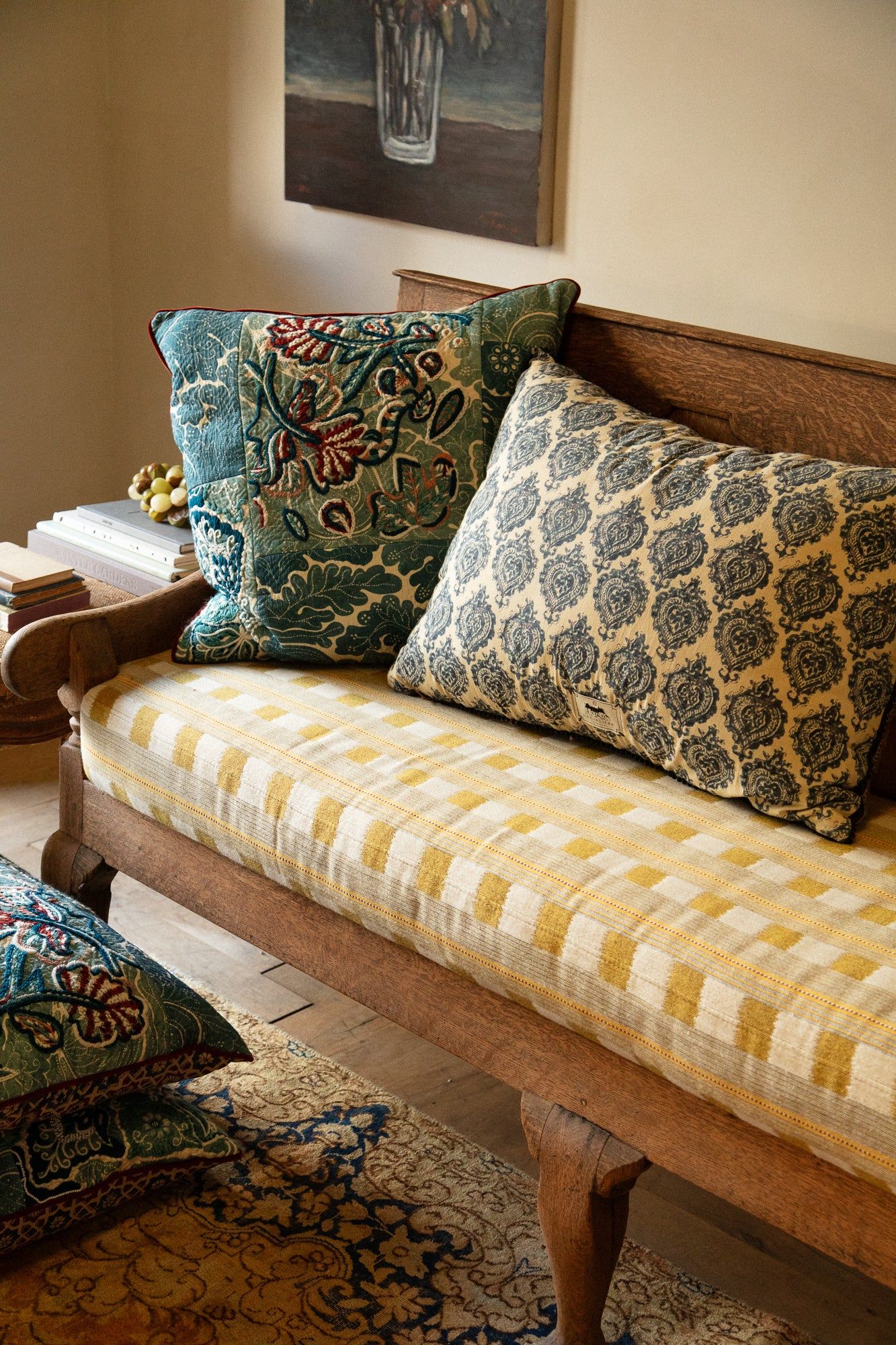 Wooden bench with yellow checkered cushion and patterned pillows. Blue floral and damask designs. Matching pillow on rug below.