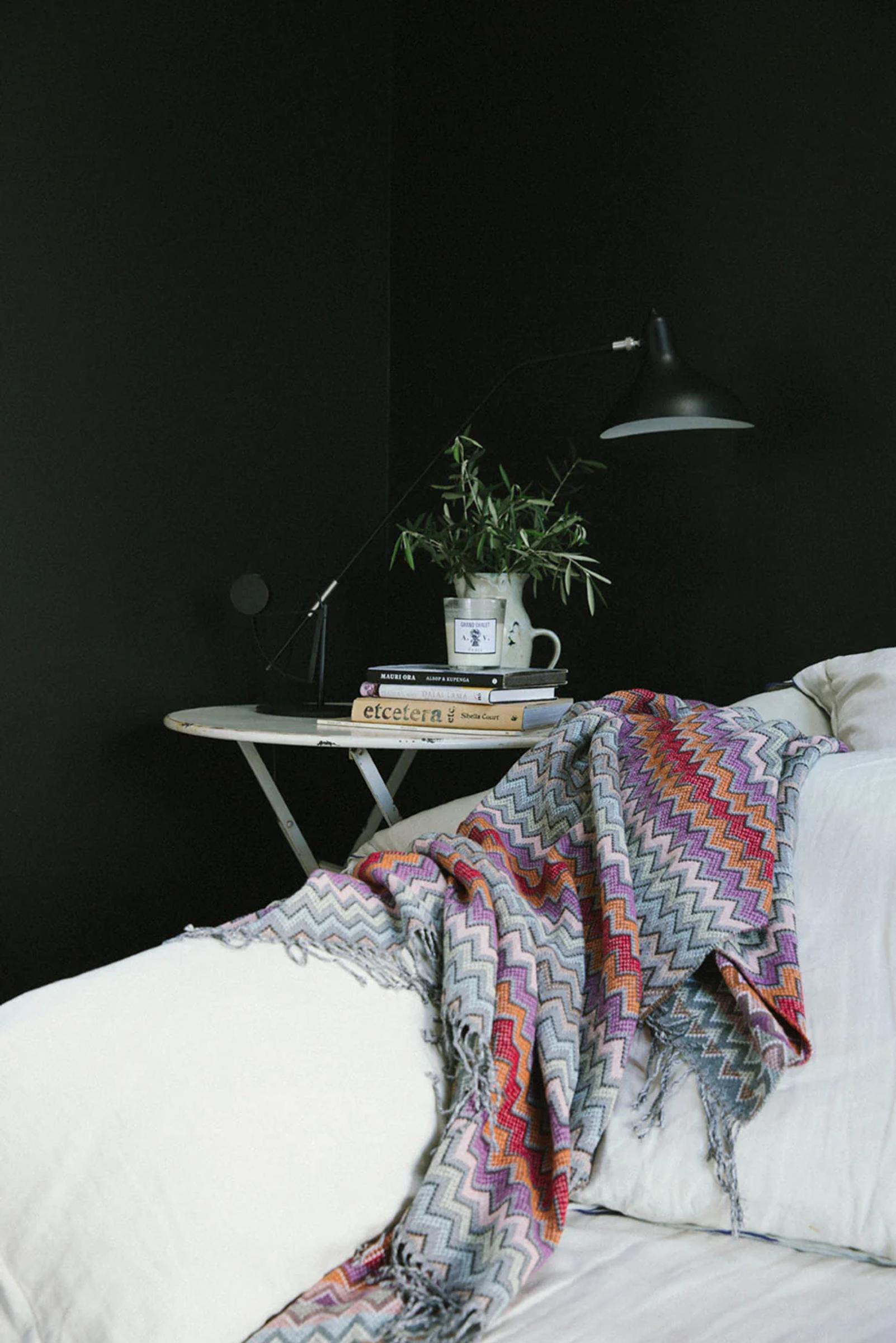 Cosy bedroom corner: Black wall, white bed with bold Missoni blanket. Round table holds plant, books, mug, and curved black lamp.