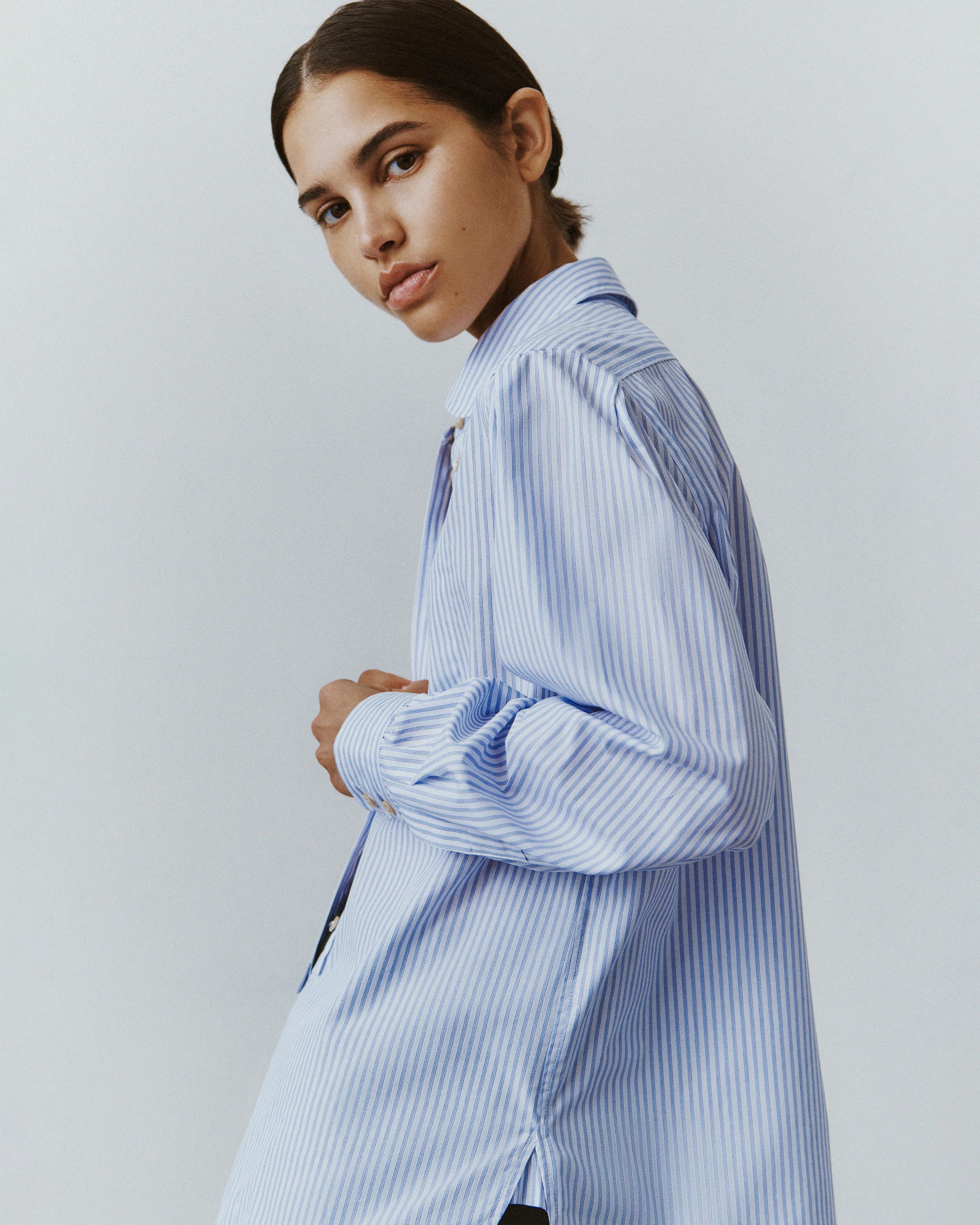 Person wearing blue and white striped button-down shirt. Dark, pulled-back hair. Light grey background.