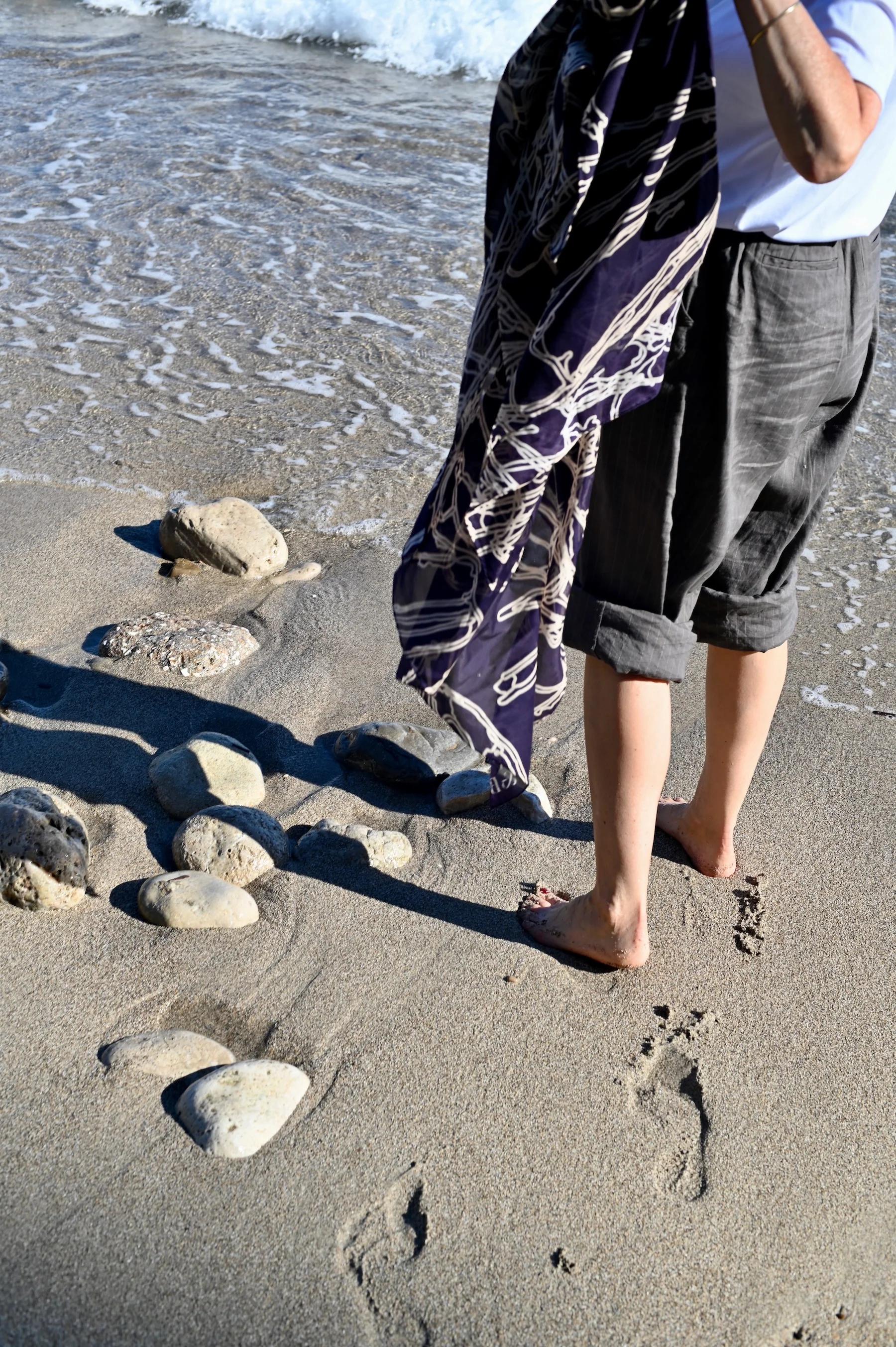 Person on sandy beach near shoreline. Holding large scarf, wearing rolled-up shorts. Footprints and smooth stones visible on sand.