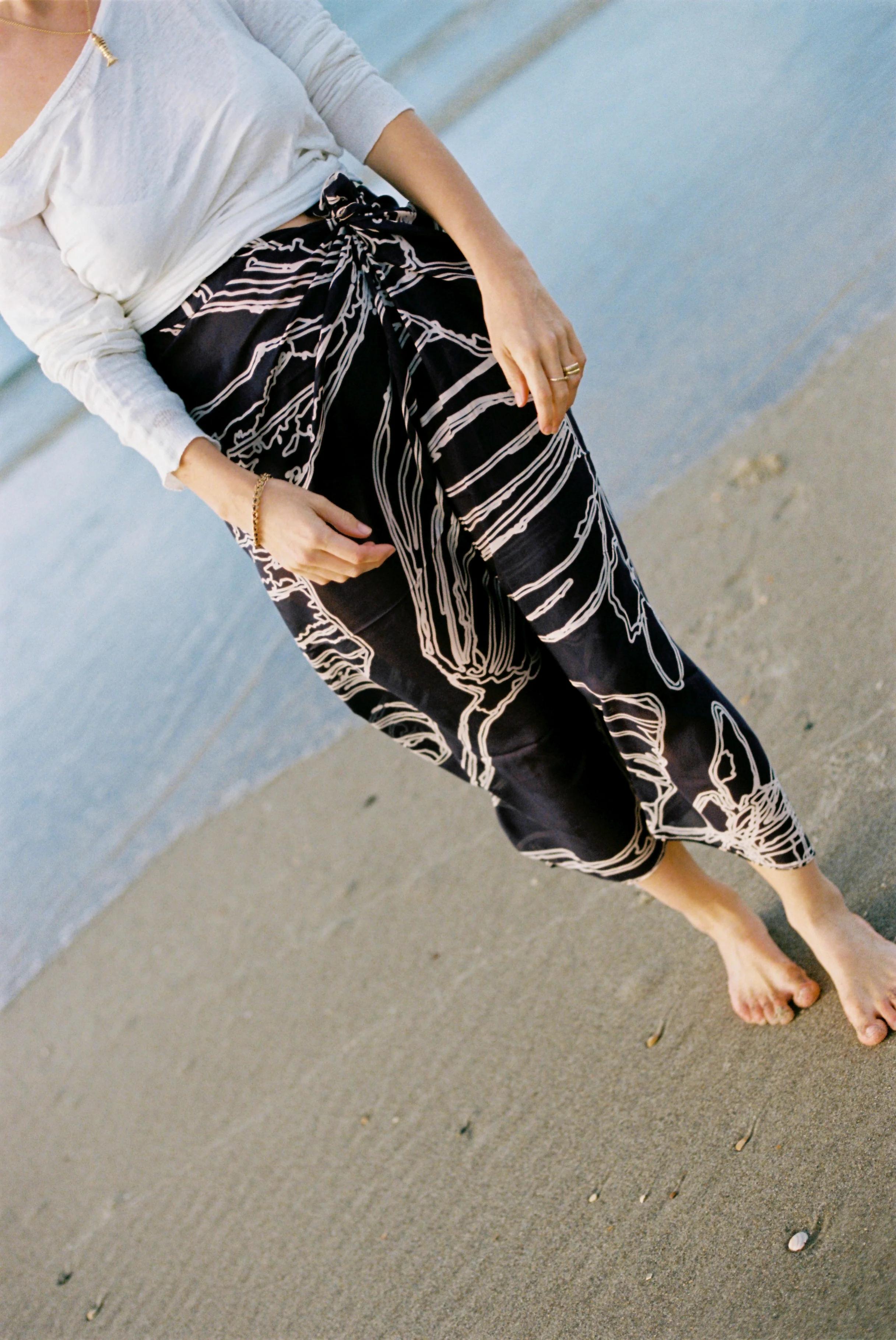 Person in white long-sleeve top and patterned black pants on sandy beach. Partially visible from shoulders to knees.