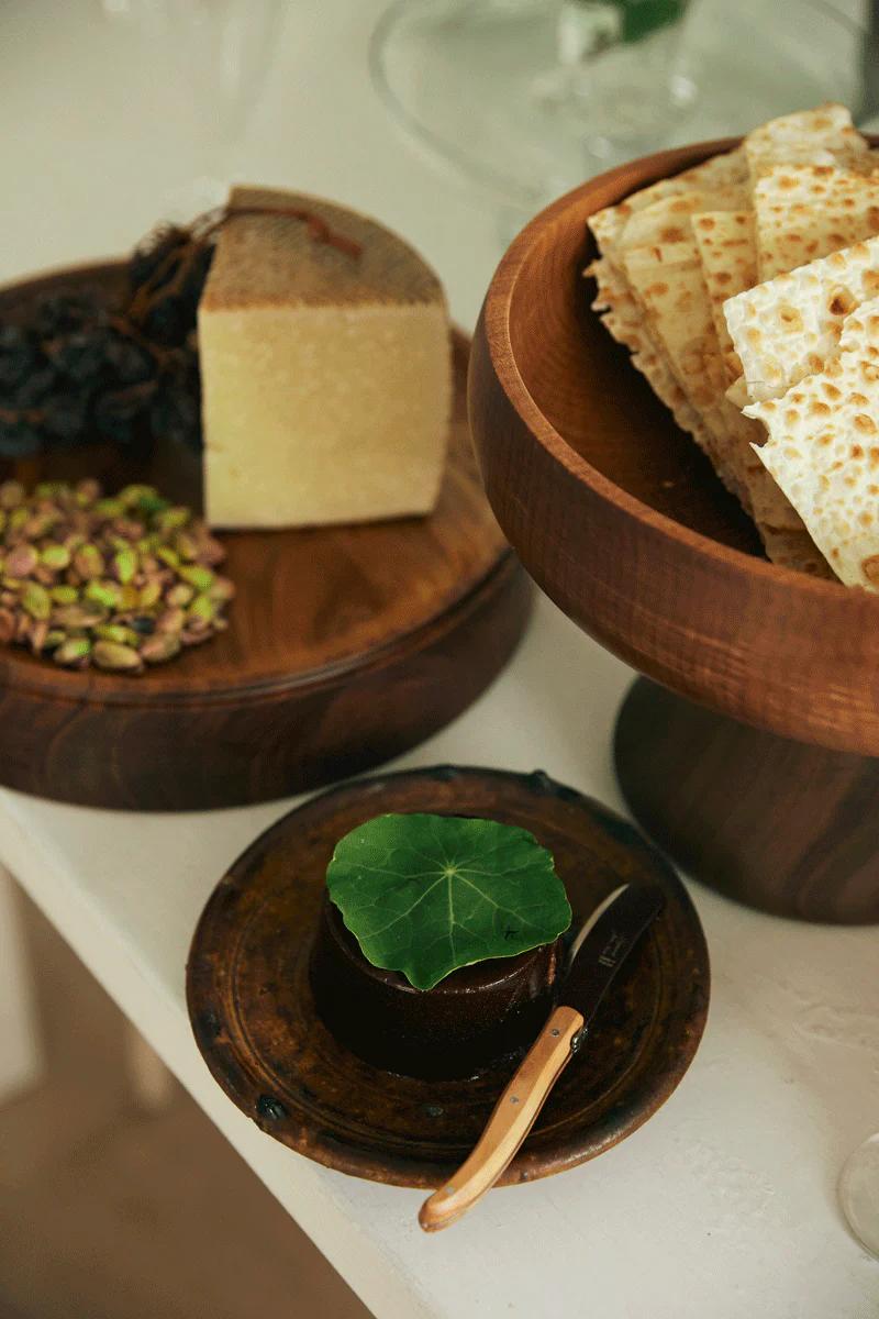 Wooden platter with cheese, grapes, and pistachios. Bowl of flatbreads and Christmas cake on round plate.