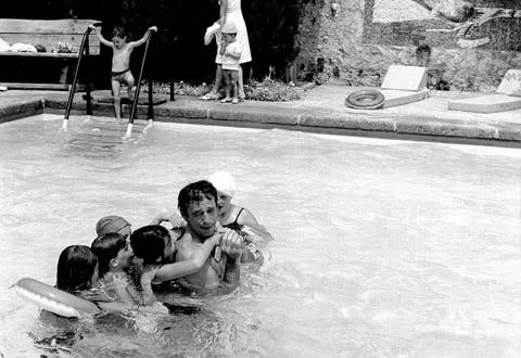 Children gathered around man in swimming pool. Child at pool edge, adult watching from deck.