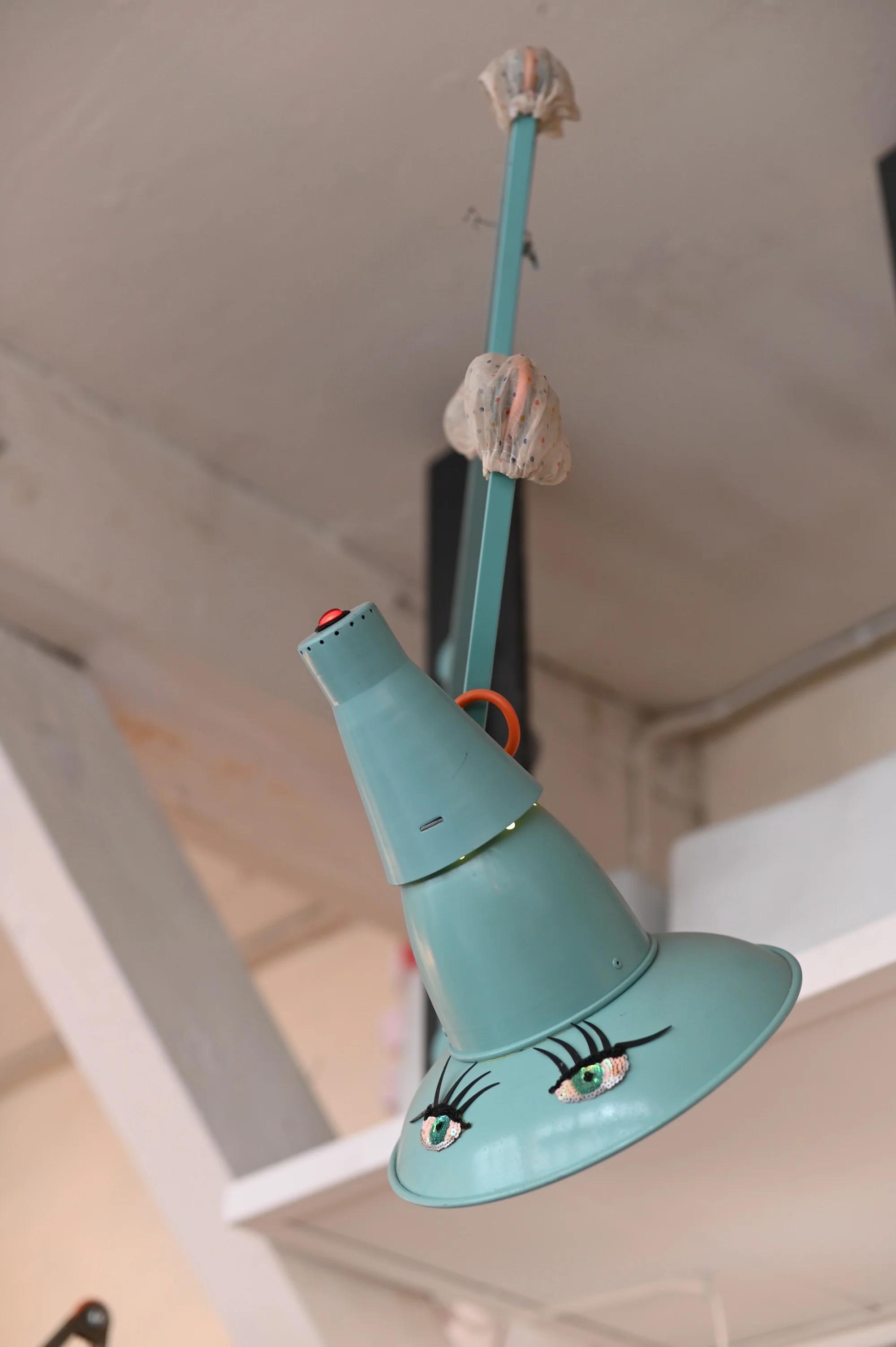 Teal cone-shaped lamp with painted eyelashes hanging from ceiling. Fabric-wrapped details at connection points.