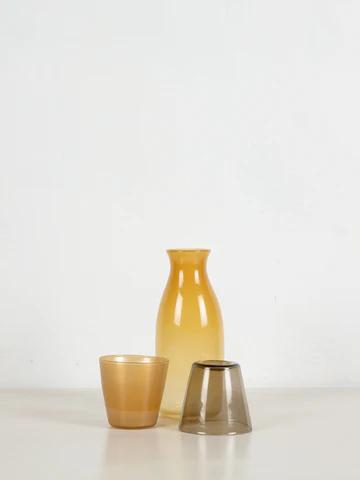 Minimalist still life: Amber vase, amber tumbler, and grey inverted cup. Translucent glass on white surface suggests sun-filled summer afternoons.