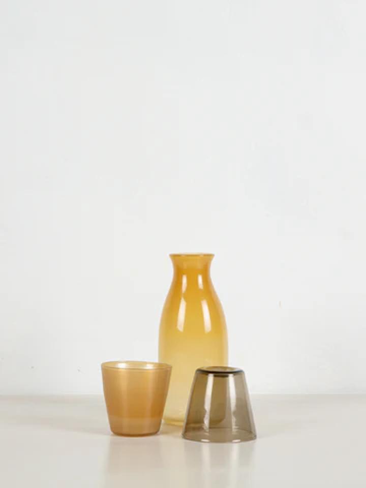 Minimalist still life: Amber vase, amber tumbler, and grey inverted cup. Translucent glass on white surface suggests sun-filled summer afternoons.