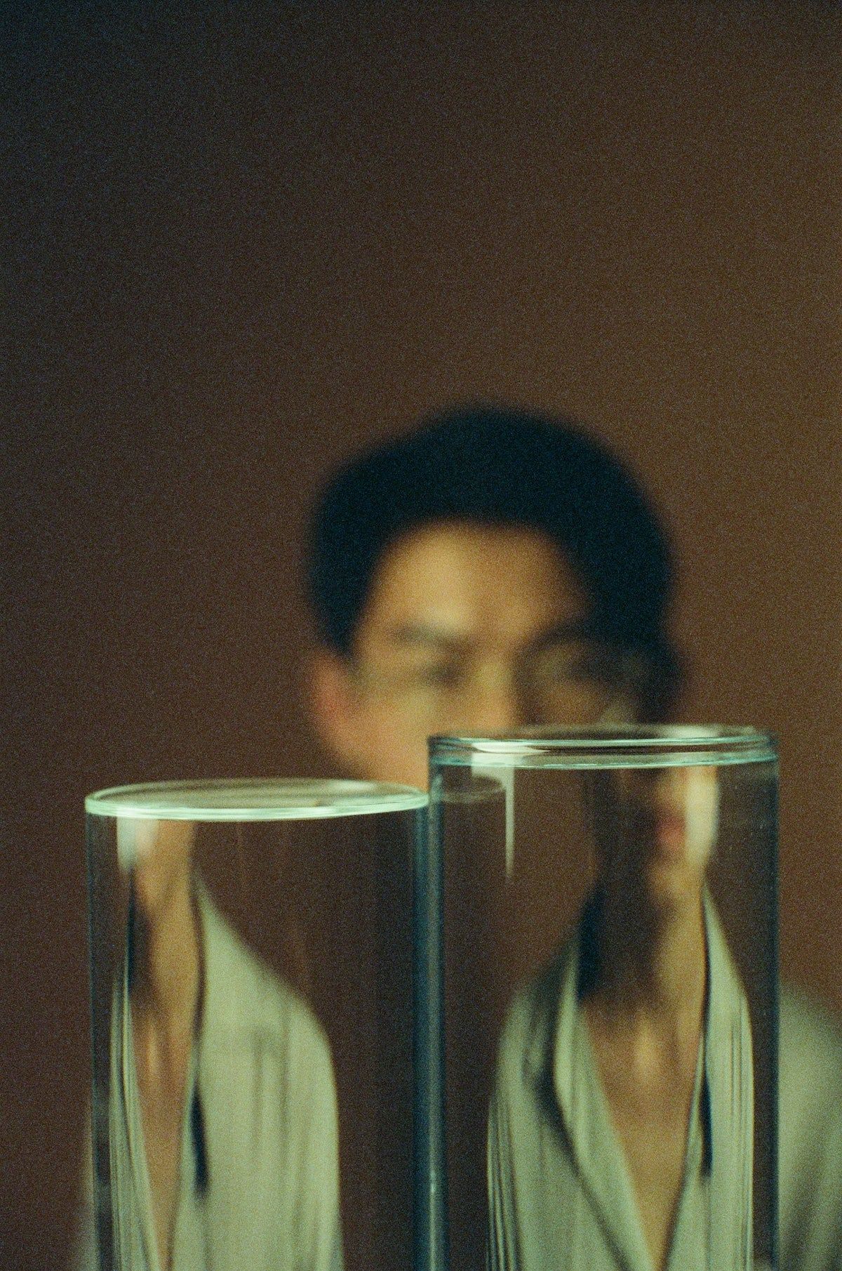 Blurry figure behind two tall, clear water-filled glass cylinders. Person's reflection visible on glass surfaces.