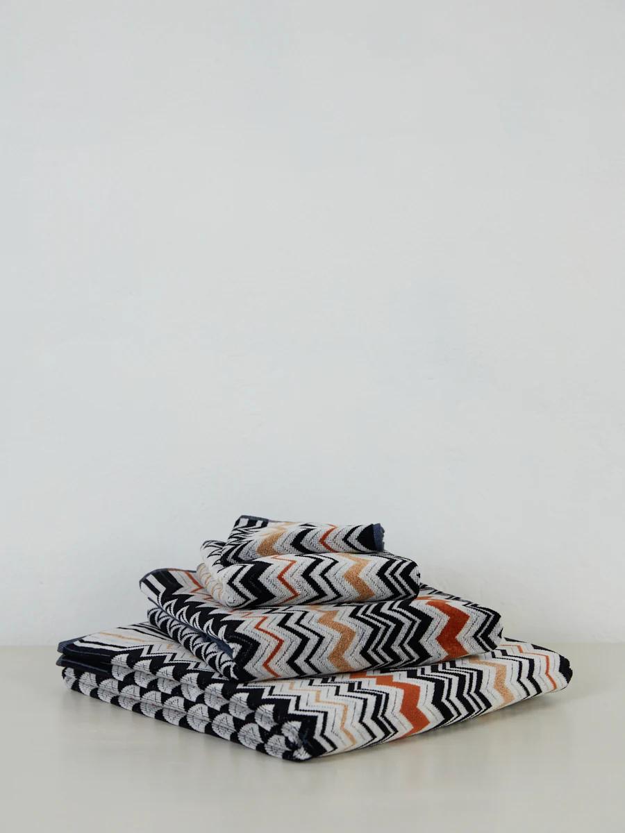 Stacked Missoni velour towels with a zigzag pattern in black, white, orange, and beige. The luxurious set sits on a light surface against a pale wall.