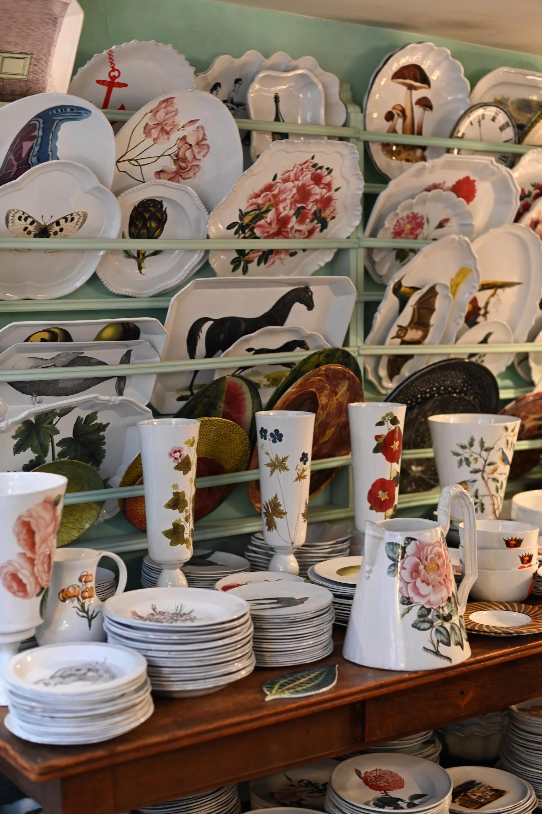 Display shelf with ceramic plates and vases. Floral, animal, and insect designs. Metal racks and wooden surface. Mint green wall.