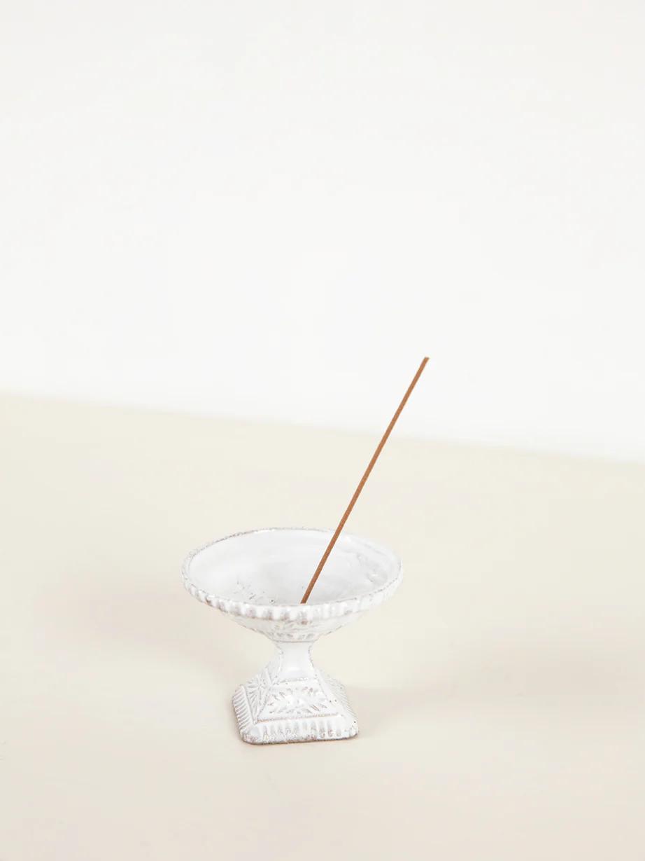 Single incense stick burning in white ceramic holder on cream-coloured surface.