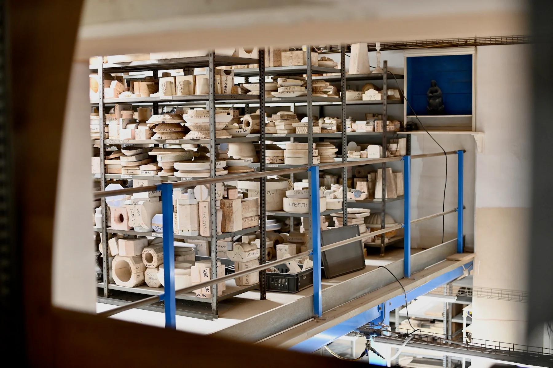 Pottery studio with molds and ceramics on metal shelves, viewed through window or opening. Laptop on lower shelf, blue door in background.