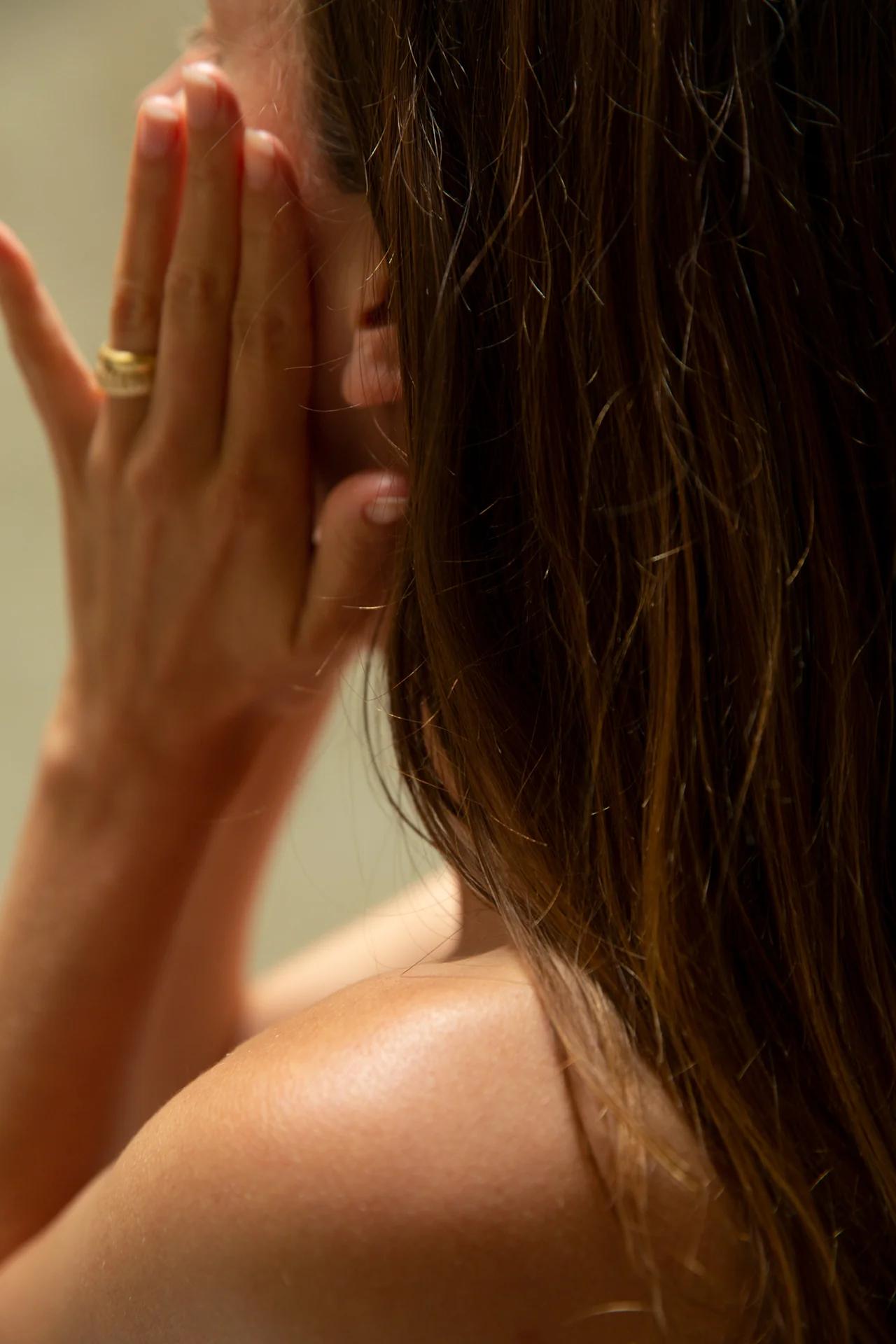 A person covers their face with both hands, a gold ring visible. The image focuses on hands and upper back, illuminated by natural light.