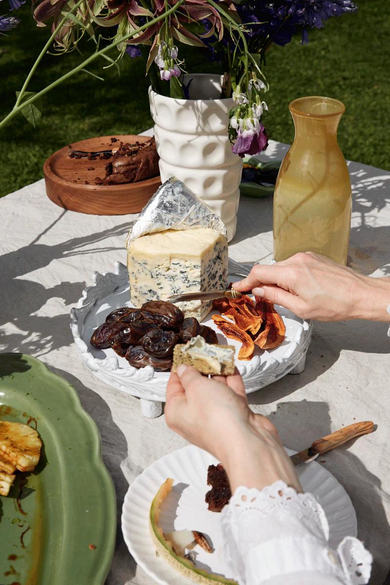 A hand spreads cheese on a cracker at an outdoor table with blue cheese, dates, and a yellow glass pitcher. Flowers add an elegant touch.