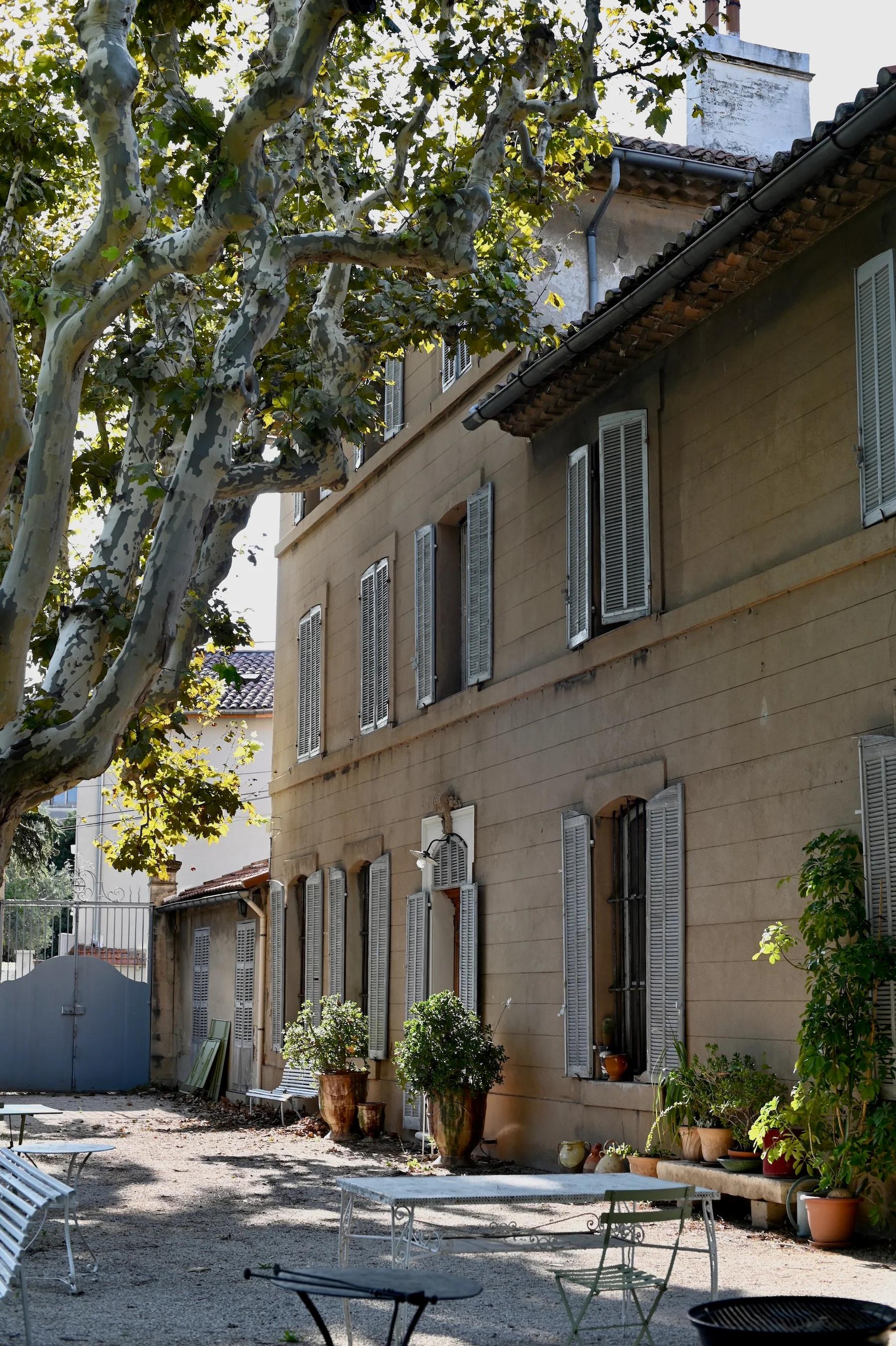 Rustic beige building with shutters under leafy trees. Outdoor patio features potted plants and metal furniture, bathed in warm, dappled sunlight.