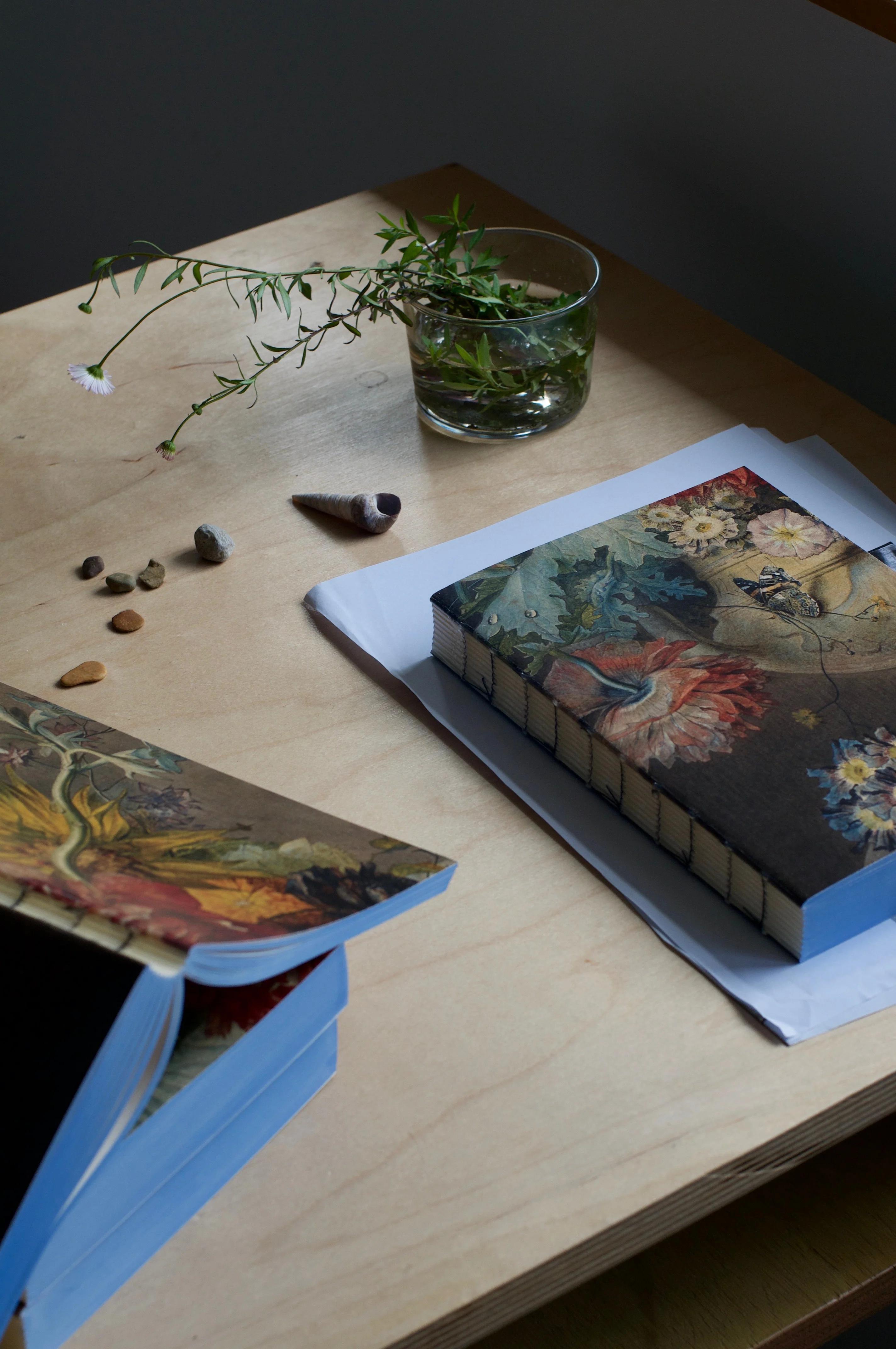 Two hardcover books with floral covers on wooden table. White sheet, notebook, glass jar with plant stems, rocks, and seashell arranged decoratively.