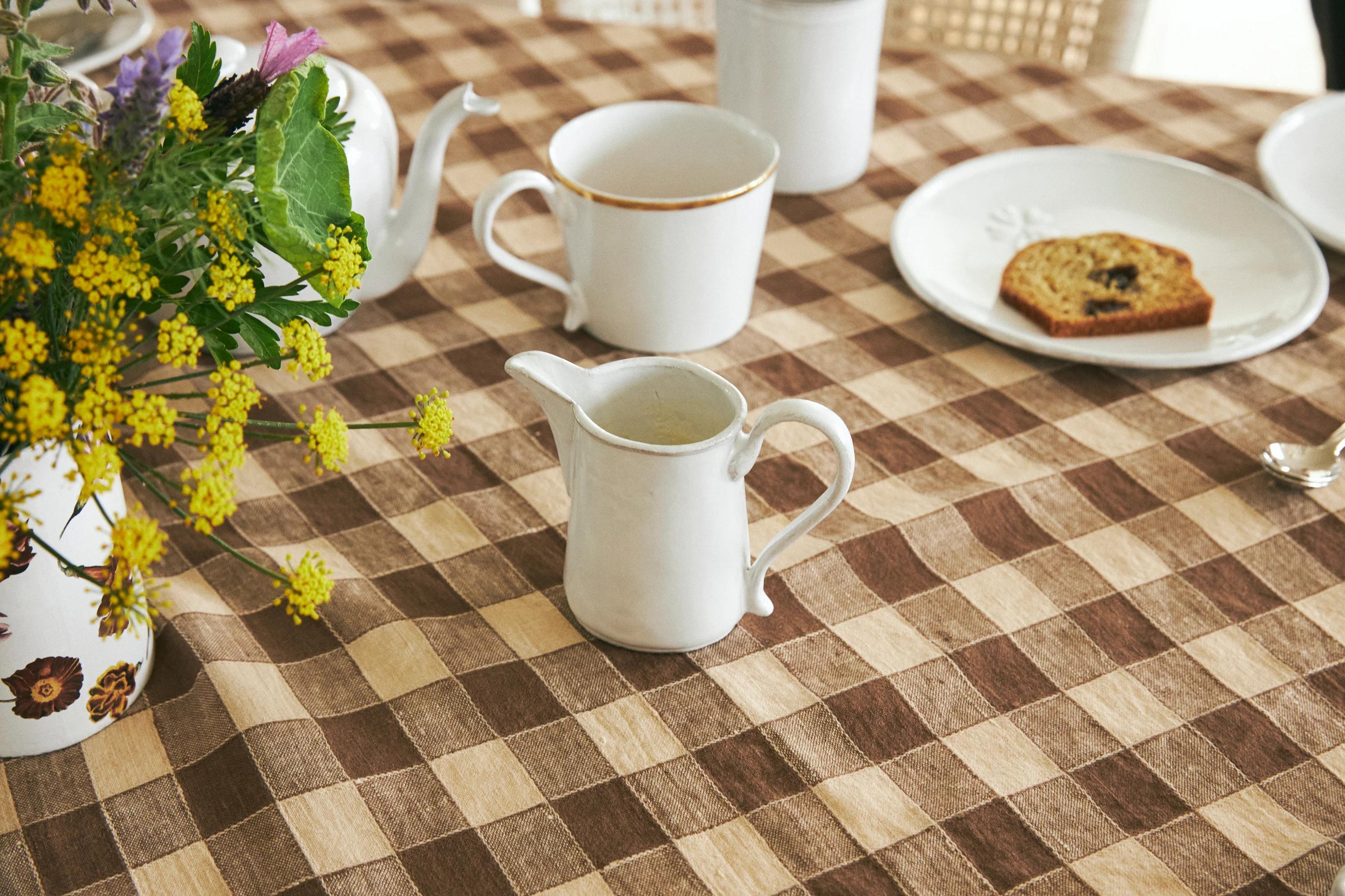 Checkered tablecloth with white pitcher, teacups, saucers. Cake slice and flower vase present.