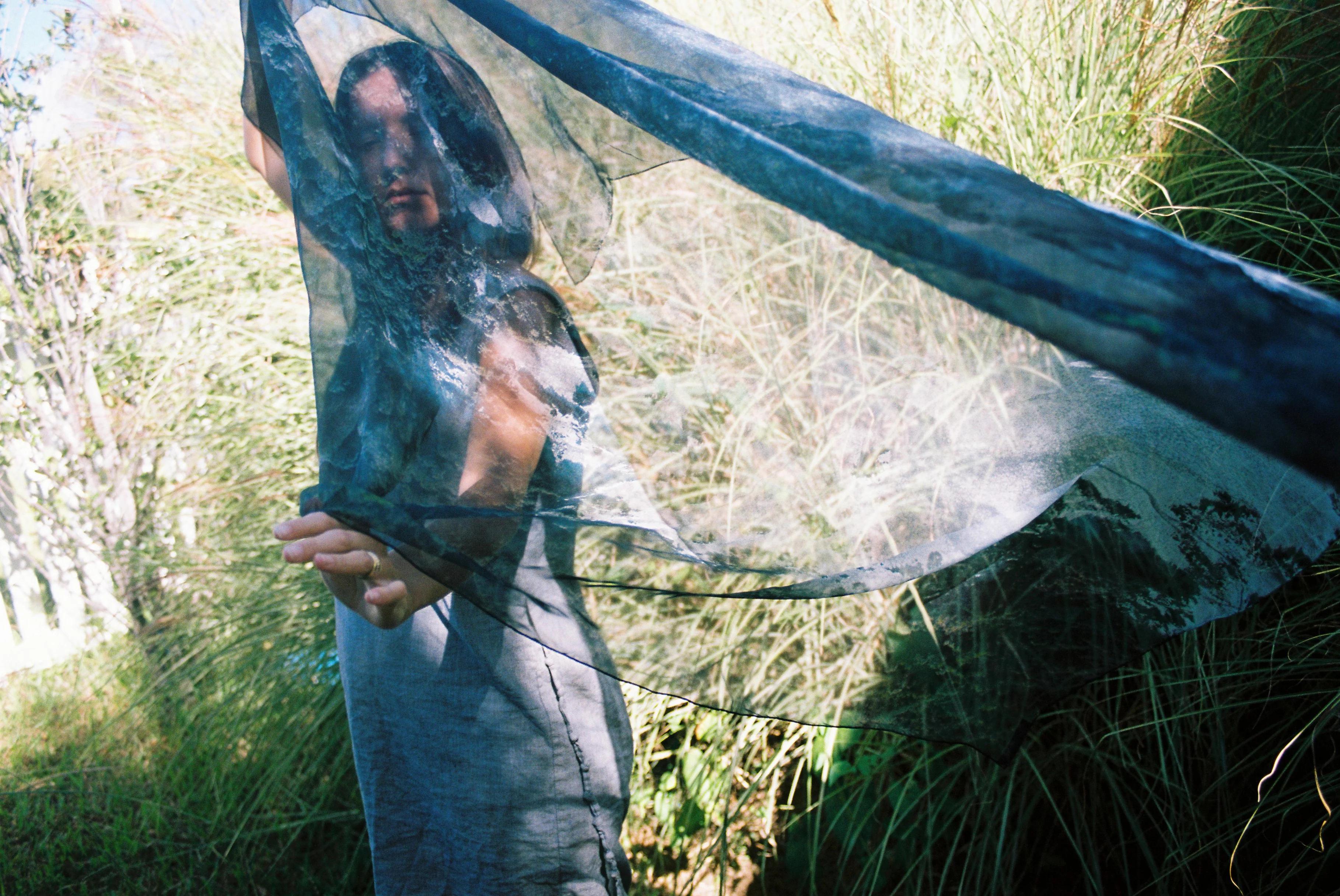 Long-haired person in flowing, semi-sheer scarf among tall grass. Sunlight catches fabric, creating ethereal effect.