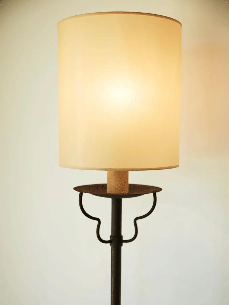 Lit Balthus Lamp with cylindrical shade. Slender metallic base with curved design. Softly lit background.