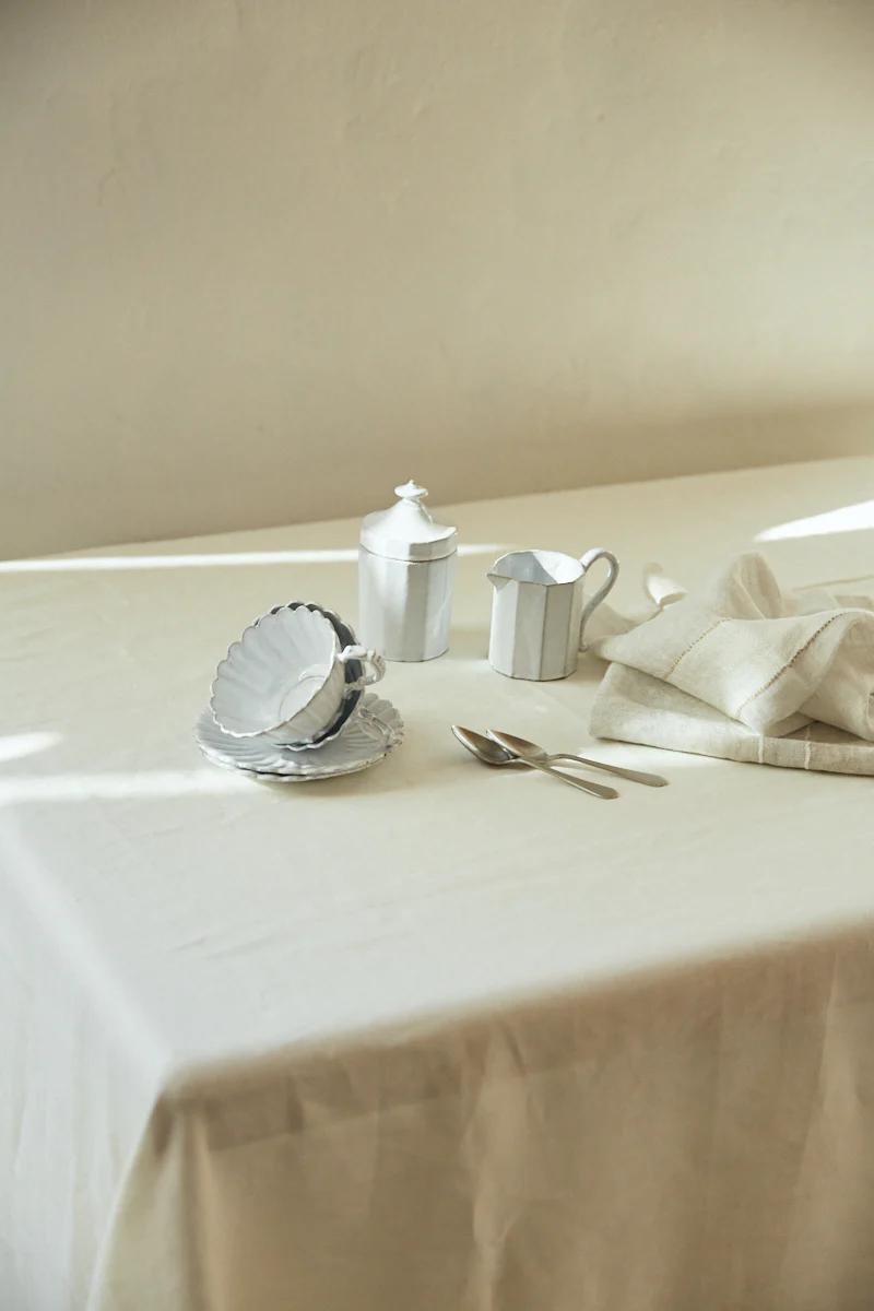 A serene tabletop with shell-shaped plates, silver spoons, a sugar bowl, and creamer on a cream tablecloth. Natural light enhances the scene.