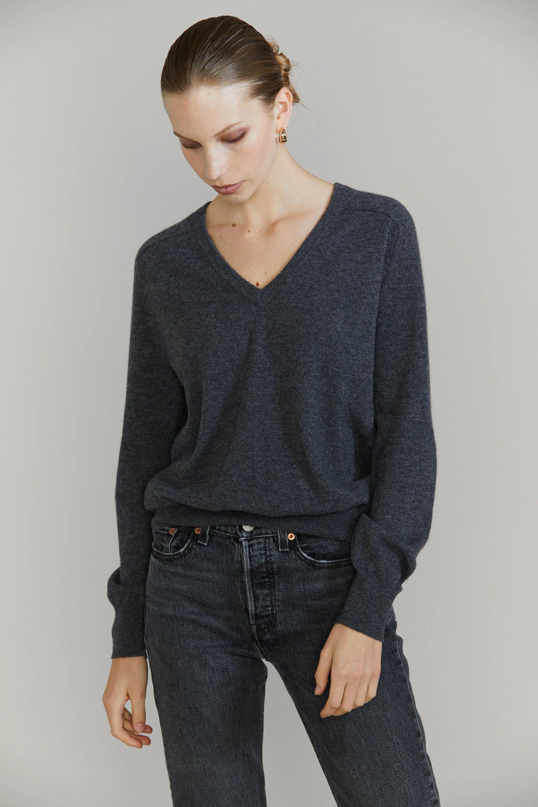 Person in dark grey V-neck sweater and jeans. Looking downward. Light-coloured background.