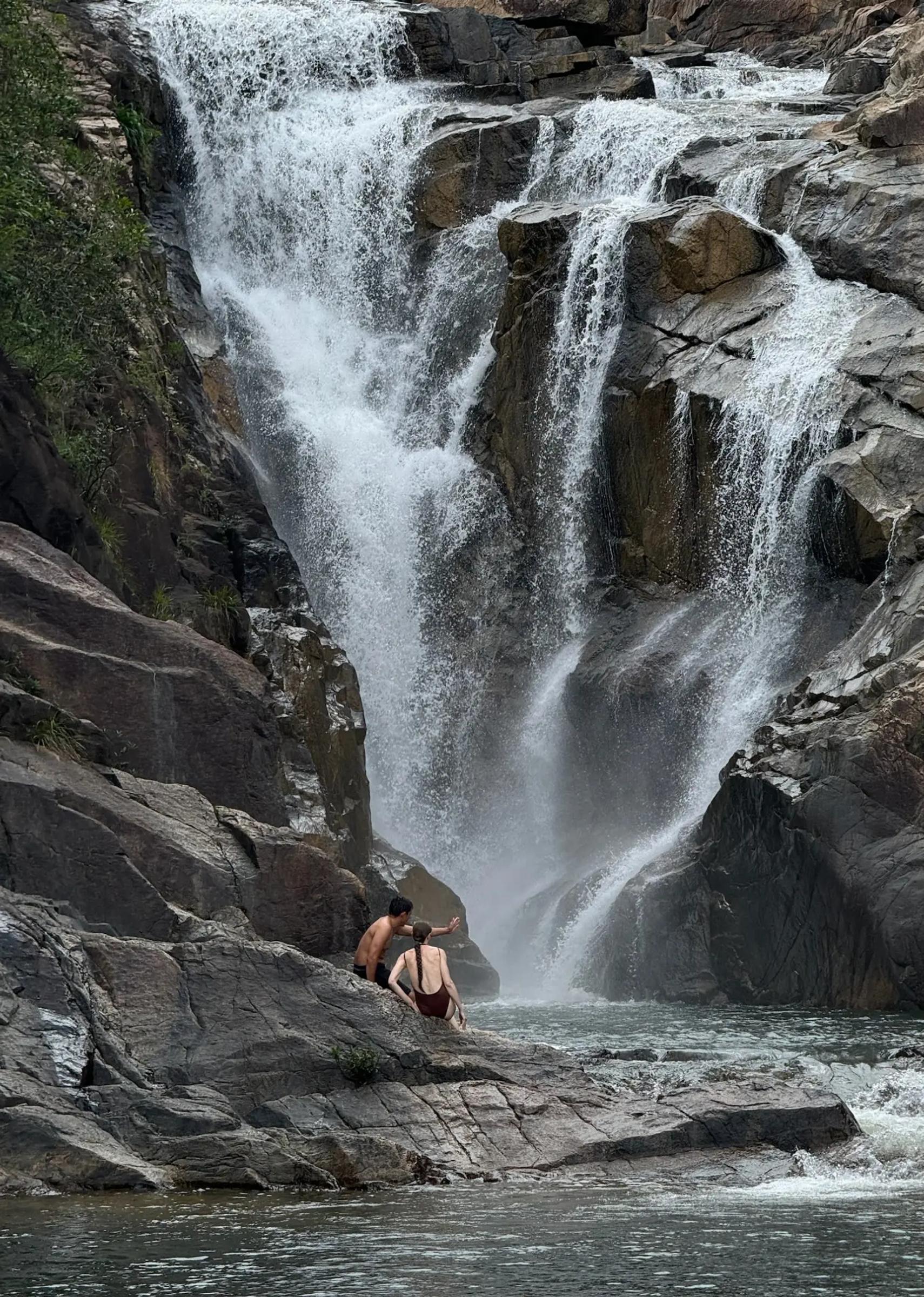 two people sit on a rock in front of a huge misty waterfall
