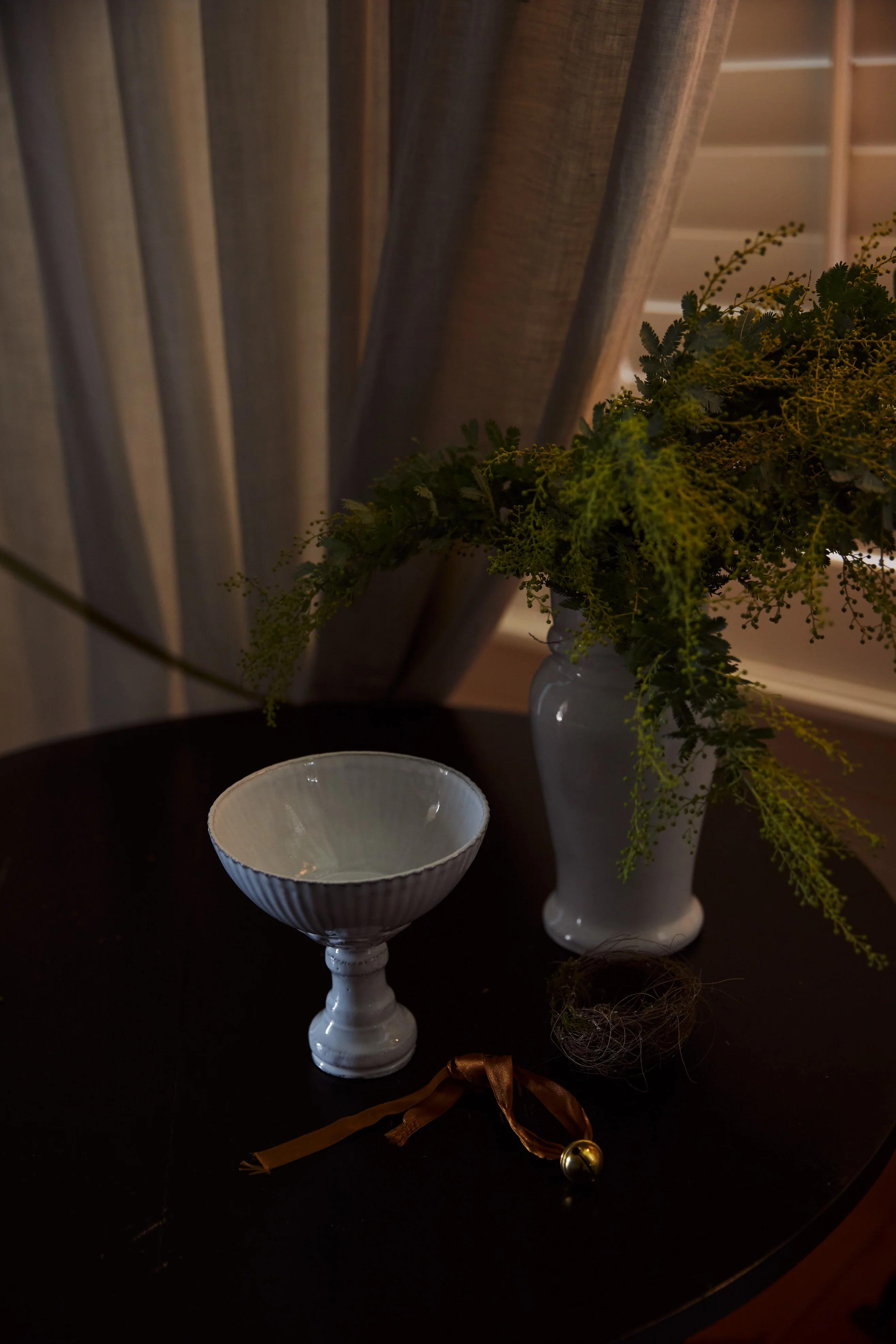 A dark wooden table holds a white ceramic bowl and vase with yellow flowers. Beige curtains partially cover a window with shutters, enhancing the bedroom’s peaceful feel.