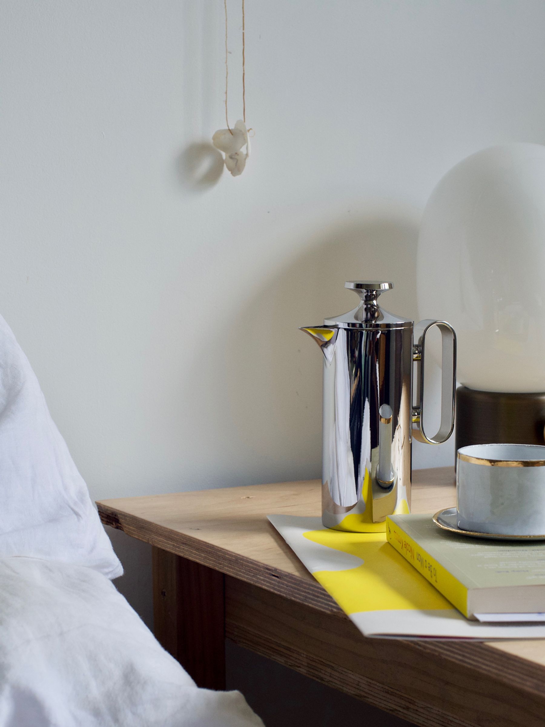 stainless steel coffee pot sits bedside next to stack of reading material and a lamp