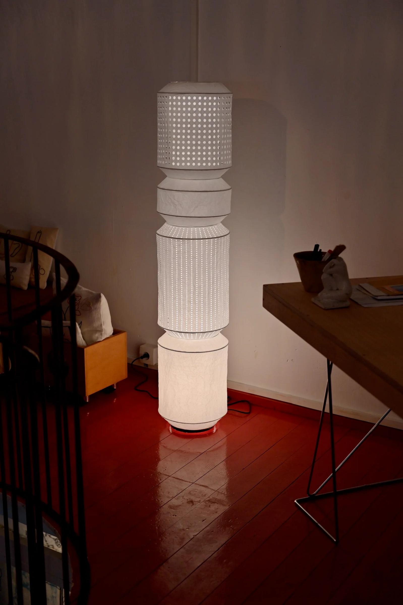 Tall lamp with intricate patterns on red floor. Spiral staircase, wooden table, and cushioned bench nearby.