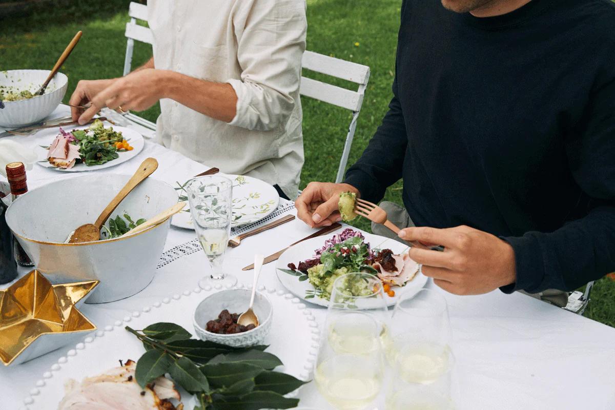 Close-up of outdoor meal table. Three people partially visible. Various dishes, drinks, and decorations on table.