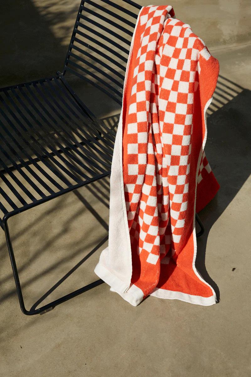 An orange and white checkered towel draped over a black metal chair on a concrete surface. Soft shadows suggest a sunny day, with the towel showcasing Baina’s mindful craftsmanship.