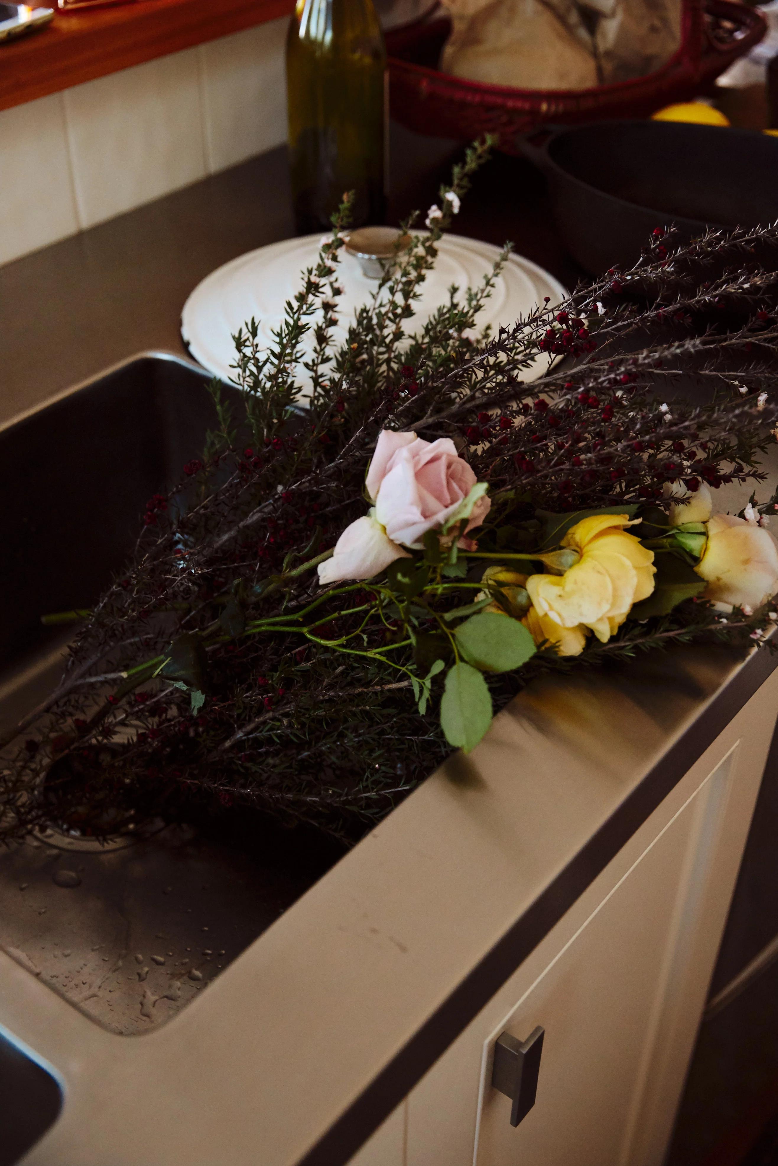 Wilting flower bouquet in sink with dishes. Cluttered countertop with green bottle and white plate.