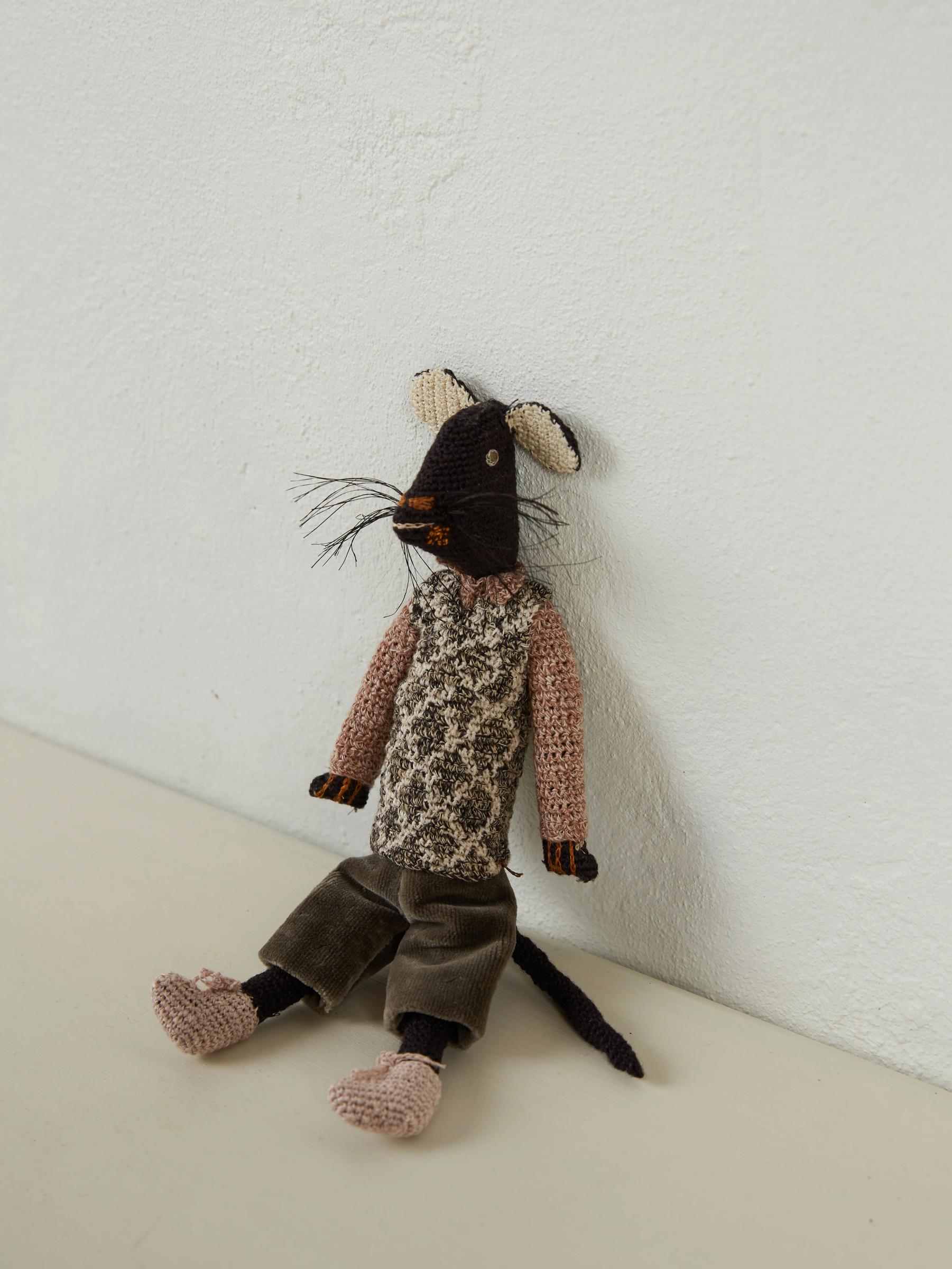 Knitted mouse doll with black and beige face, patterned shirt, and green pants. Pink limbs and string whiskers. Sits against white wall, reminiscent of Sophie Digard's work.