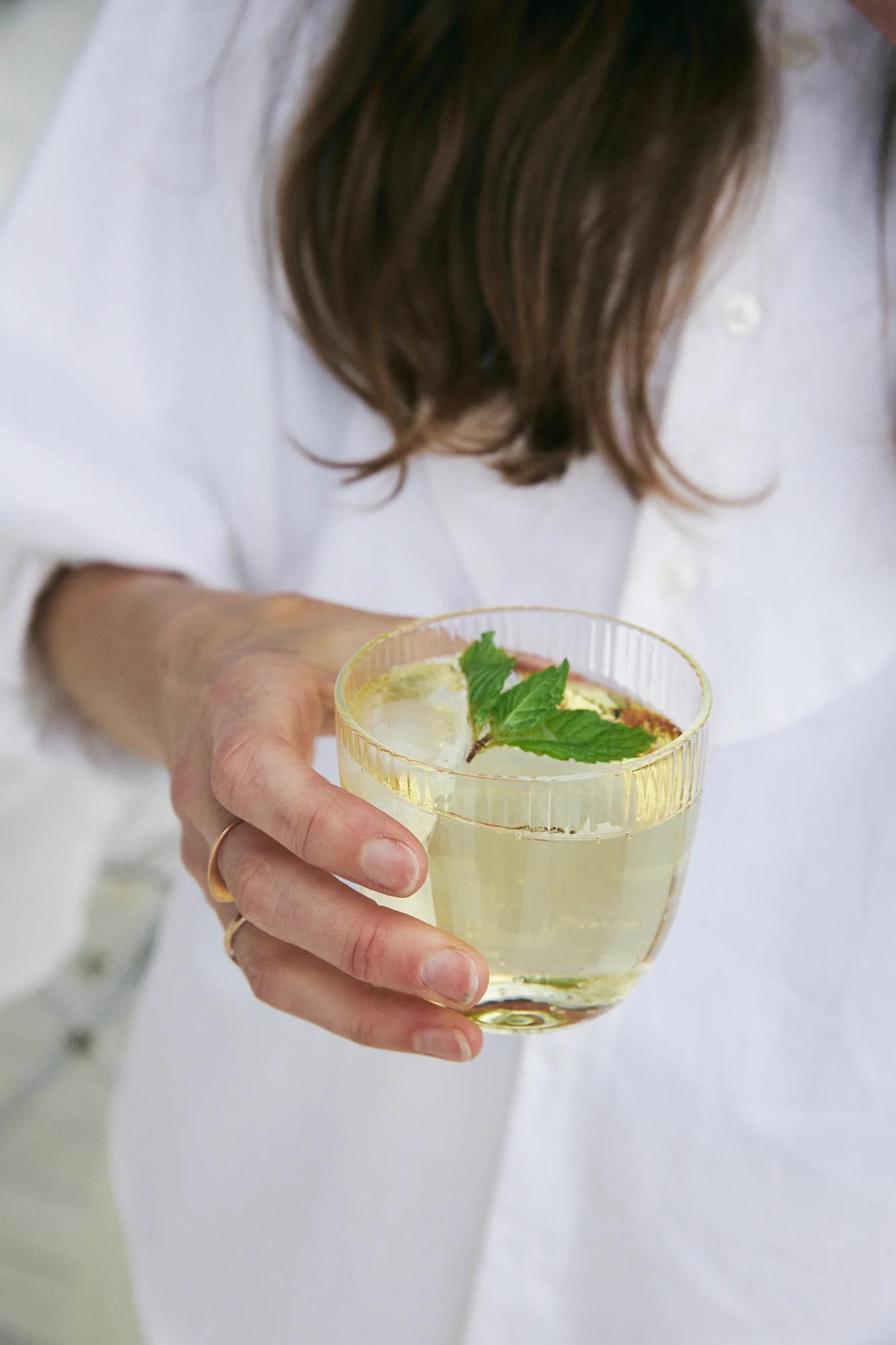 Person with long brown hair holds glass of light-coloured drink with ice and mint. Focus on hand and refreshment.