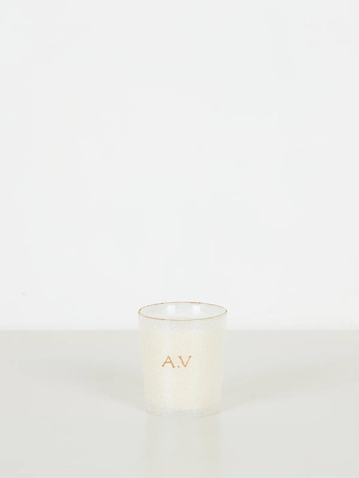 Small white candle holder with glittery finish. Gold "A.V" initials near top. Light-colored surface, white background.