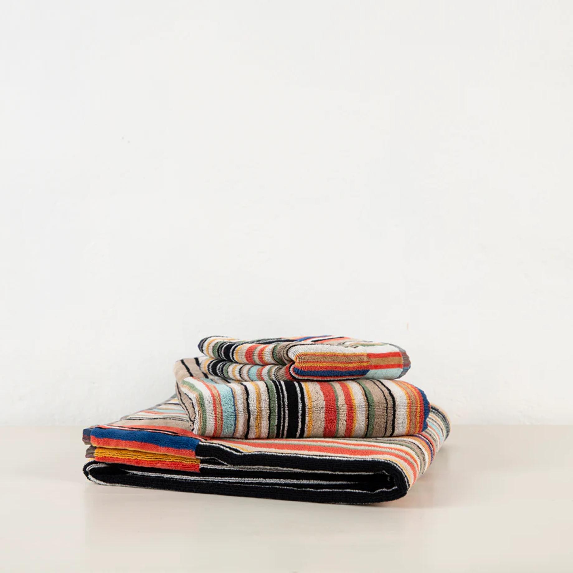 Stack of three Missoni towels with bold stripes. Colours include red, orange, blue, black, beige, and white on light background.