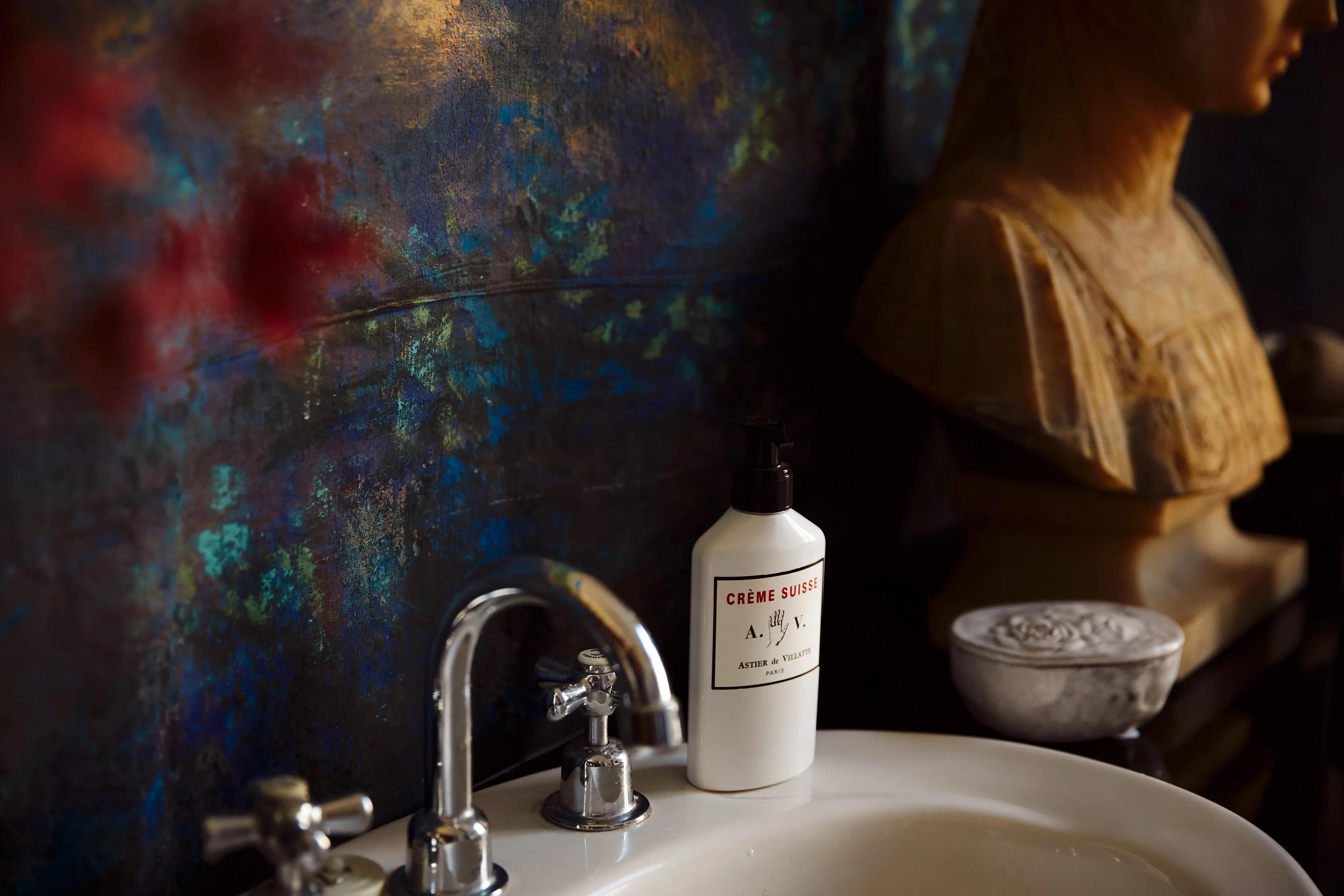 Bathroom sink with silver tap and Crème Suisse lotion. Dark, multicoloured wall and partial bust statue create artistic, winter solstice-like atmosphere.