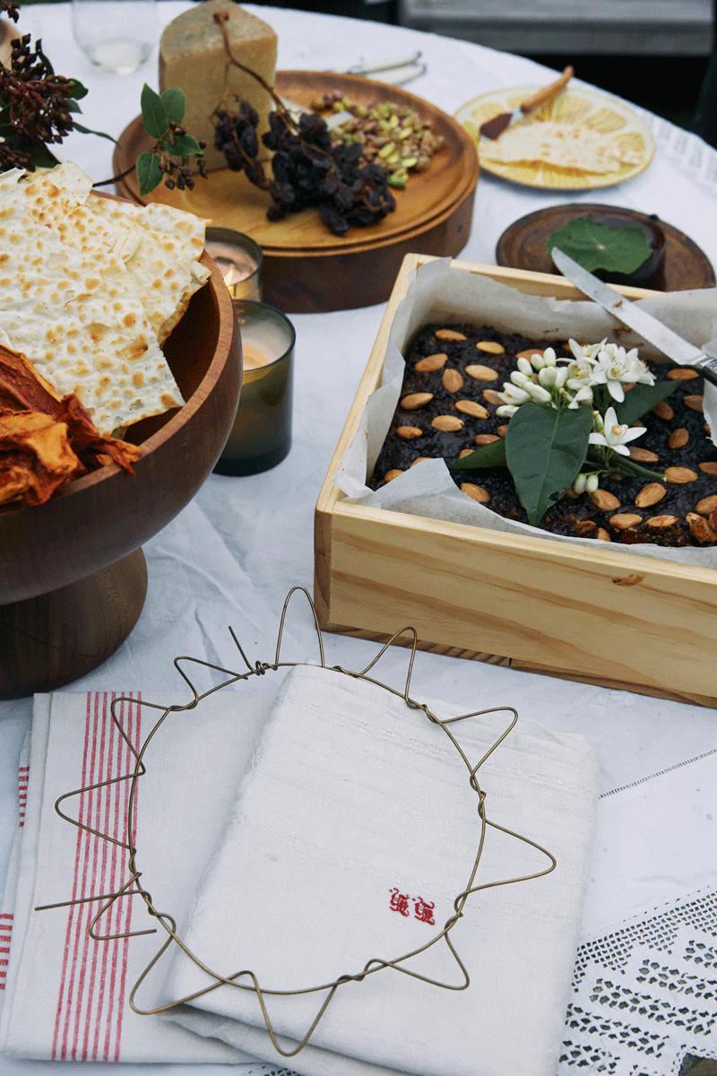 Table with flatbreads, nut-decorated dessert, cheese board, and decorative items. Folded cloths visible.