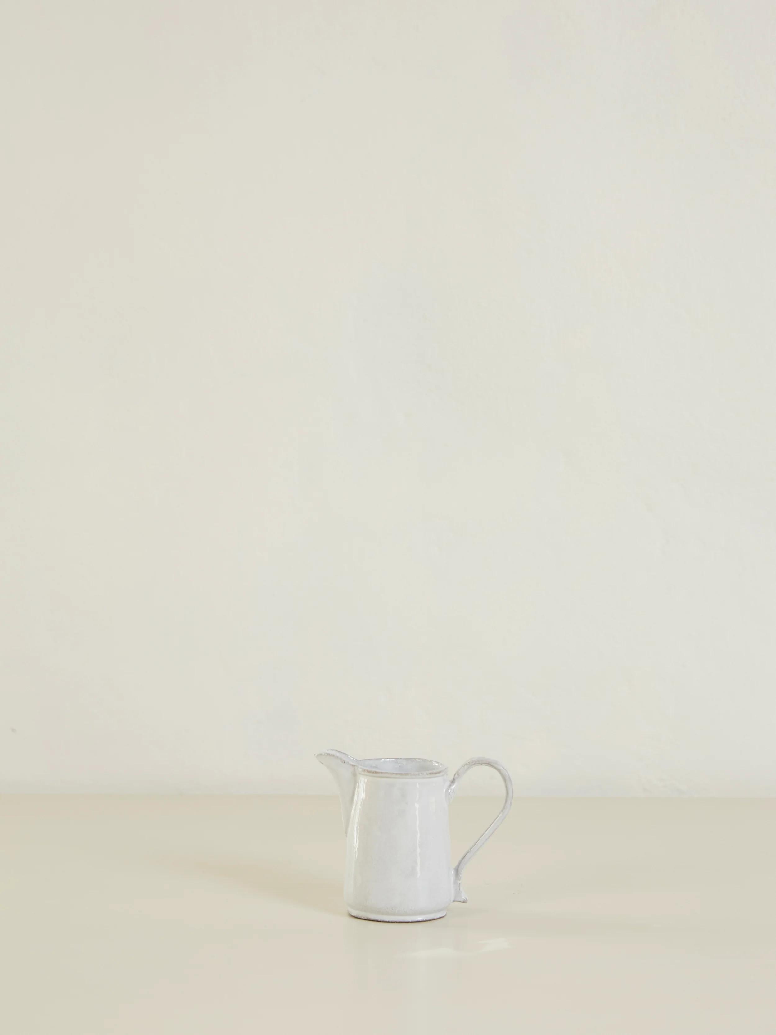 Small white ceramic pitcher on light surface. Plain, off-white background. Curved handle and narrow spout.