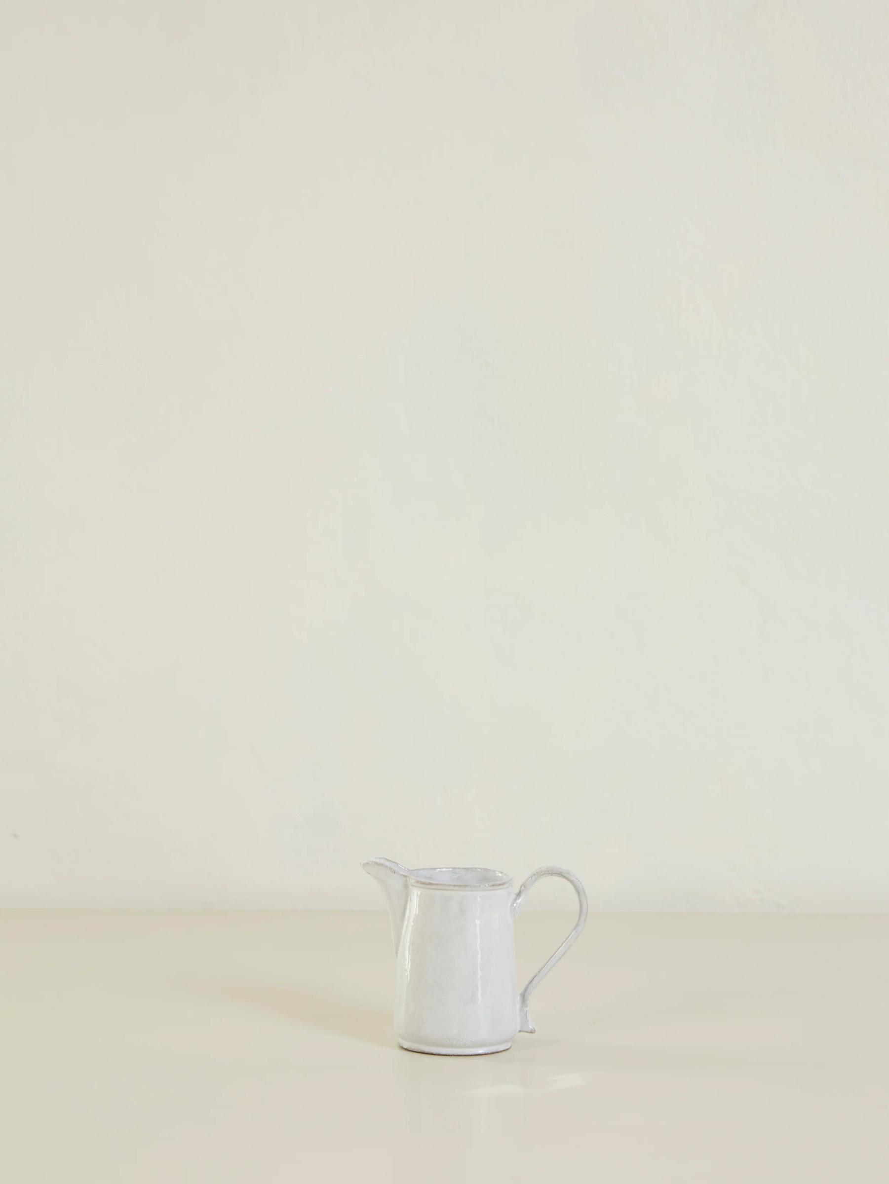 Small white ceramic pitcher on light surface. Plain, off-white background. Curved handle and narrow spout.
