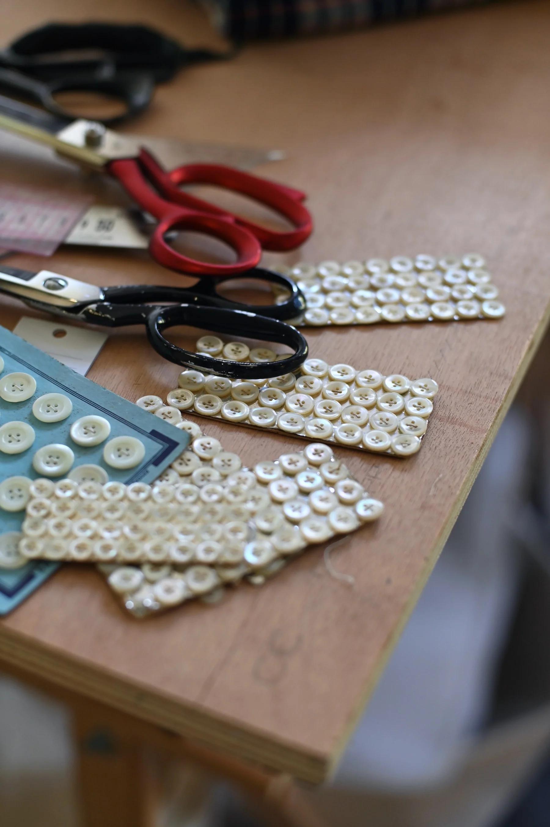 Red-handled scissors and white button cards on wooden tabletop. Fabric remnants visible.