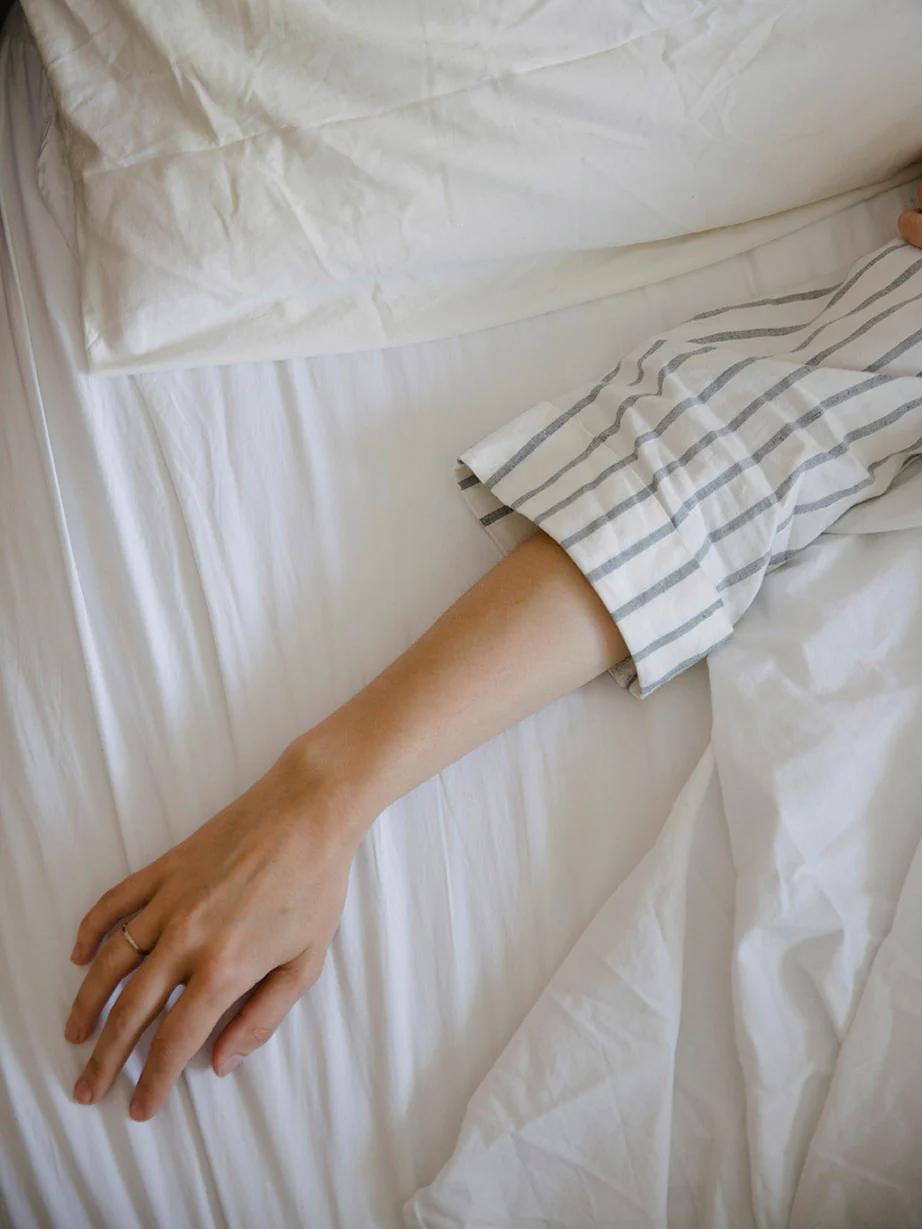 Person in striped shirt lying on white sheets. Left arm extended, simple ring on finger. Peaceful, relaxed pose.