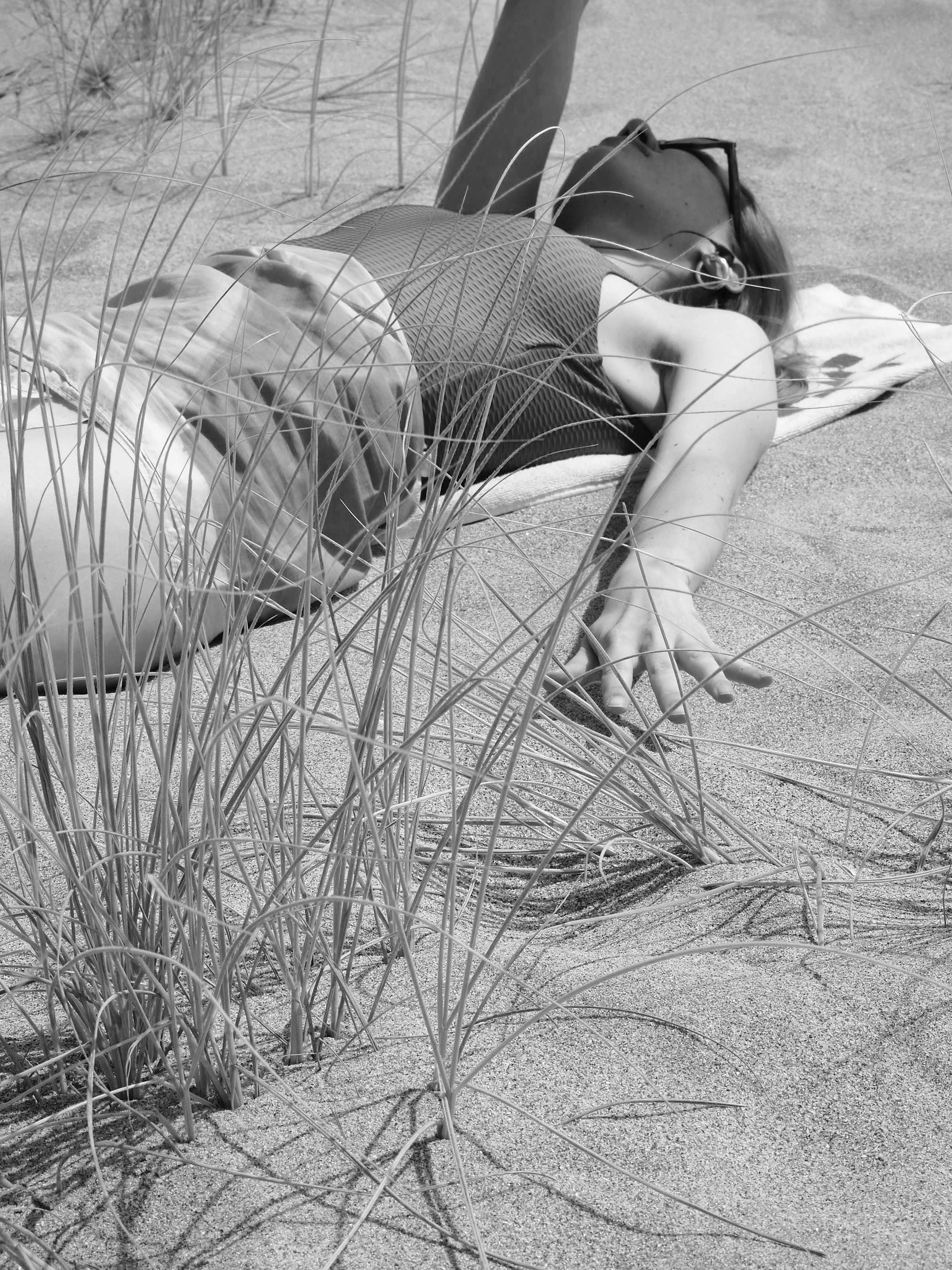 A person wearing sunglasses stretches an arm while lying on a beach towel in the sand, surrounded by sparse grass. The image evokes a relaxed summer vibe.