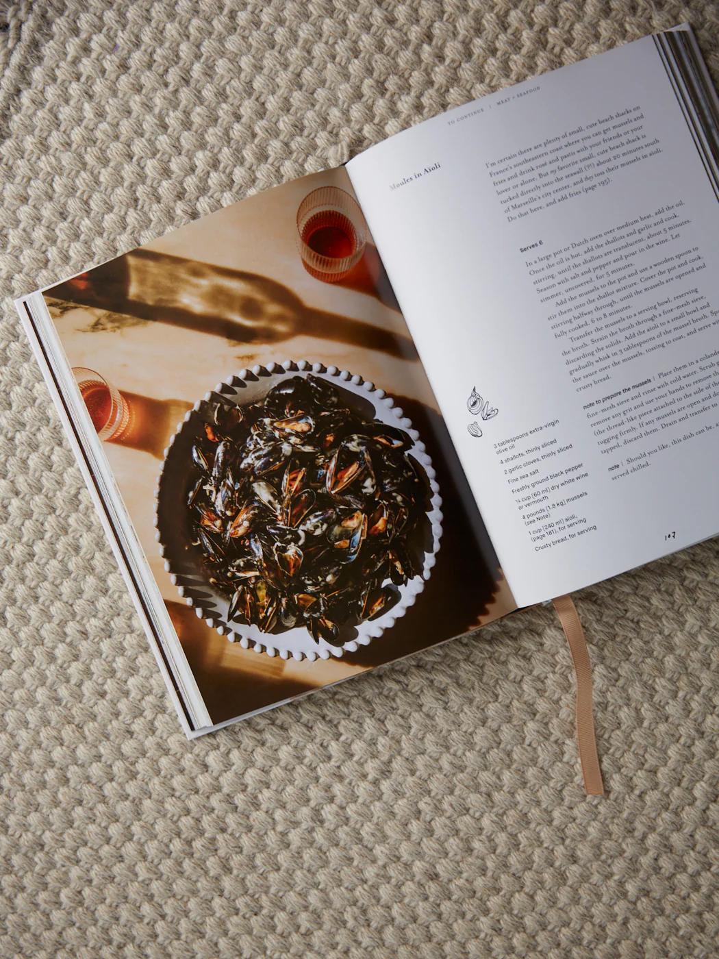 Open cookbook showing mussels dish photo and recipe. Beige ribbon bookmark visible. Textured beige surface.